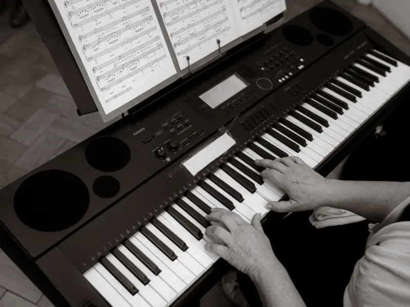 Nahaufnahme einer Person, die mit beiden Händen auf einem Casio-Keyboard spielt. Vor ihr steht ein Notenständer mit Notenblättern, auf denen Akkorde wie Fmaj7 und Em7 zu sehen sind. Die Szene vermittelt eine konzentrierte Atmosphäre beim Musizieren.