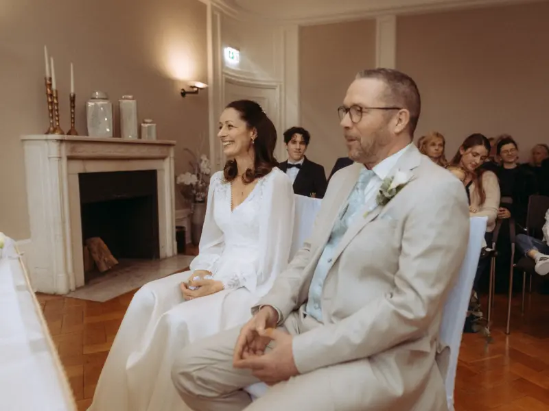 Ein glückliches Brautpaar sitzt während der Trauung in einem eleganten Saal vor einem Kamin, festgehalten von einem Hochzeitsfotograf in Schwerin und MV. Die Braut trägt ein weißes Spitzenkleid, der Bräutigam einen hellen Anzug; im Hintergrund verfolgen Gäste die Zeremonie.