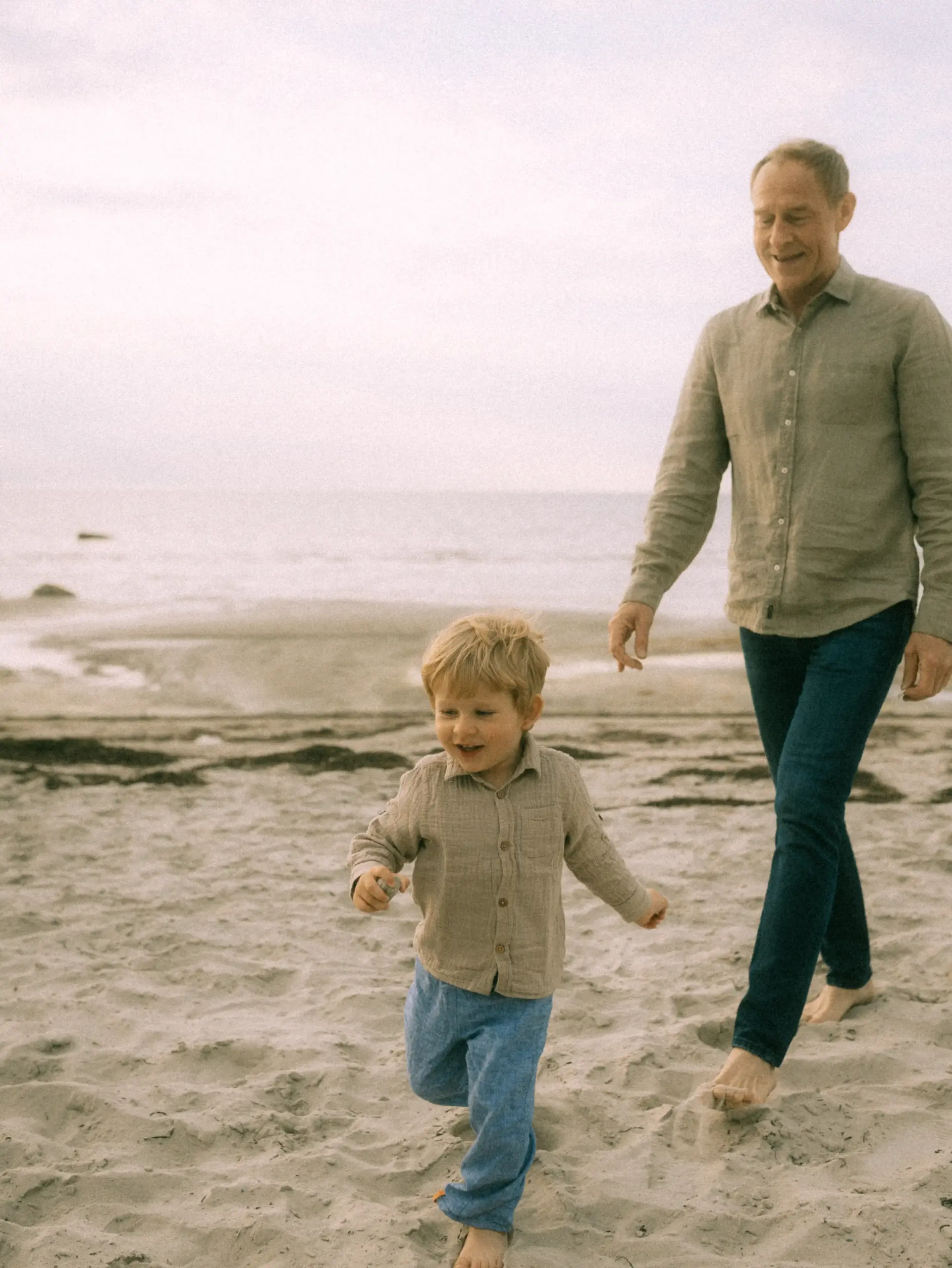 Ein kleiner Junge läuft fröhlich über den Sandstrand, während sein Vater ihn lächelnd begleitet. Die beiden tragen aufeinander abgestimmte Outfits in Naturtönen, was den entspannten Moment am Meer unterstreicht. Authentische Familienfotos in Schwerin festgehalten.