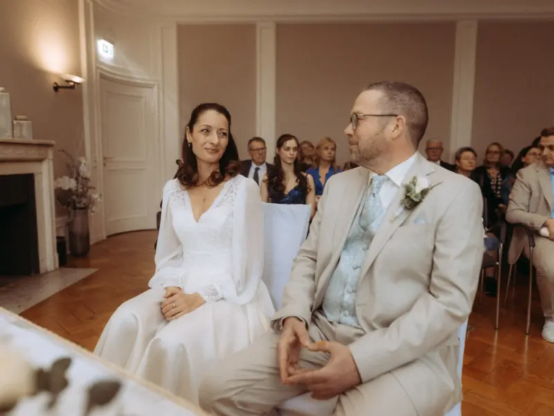 Ein glückliches Brautpaar sitzt während der Trauung in einem eleganten Saal, aufgenommen vom Hochzeitsfotograf in Schwerin und MV. Die Braut trägt ein weißes Langarmkleid und lächelt den Bräutigam im hellen Anzug an, während Gäste im Hintergrund die Zeremonie verfolgen.