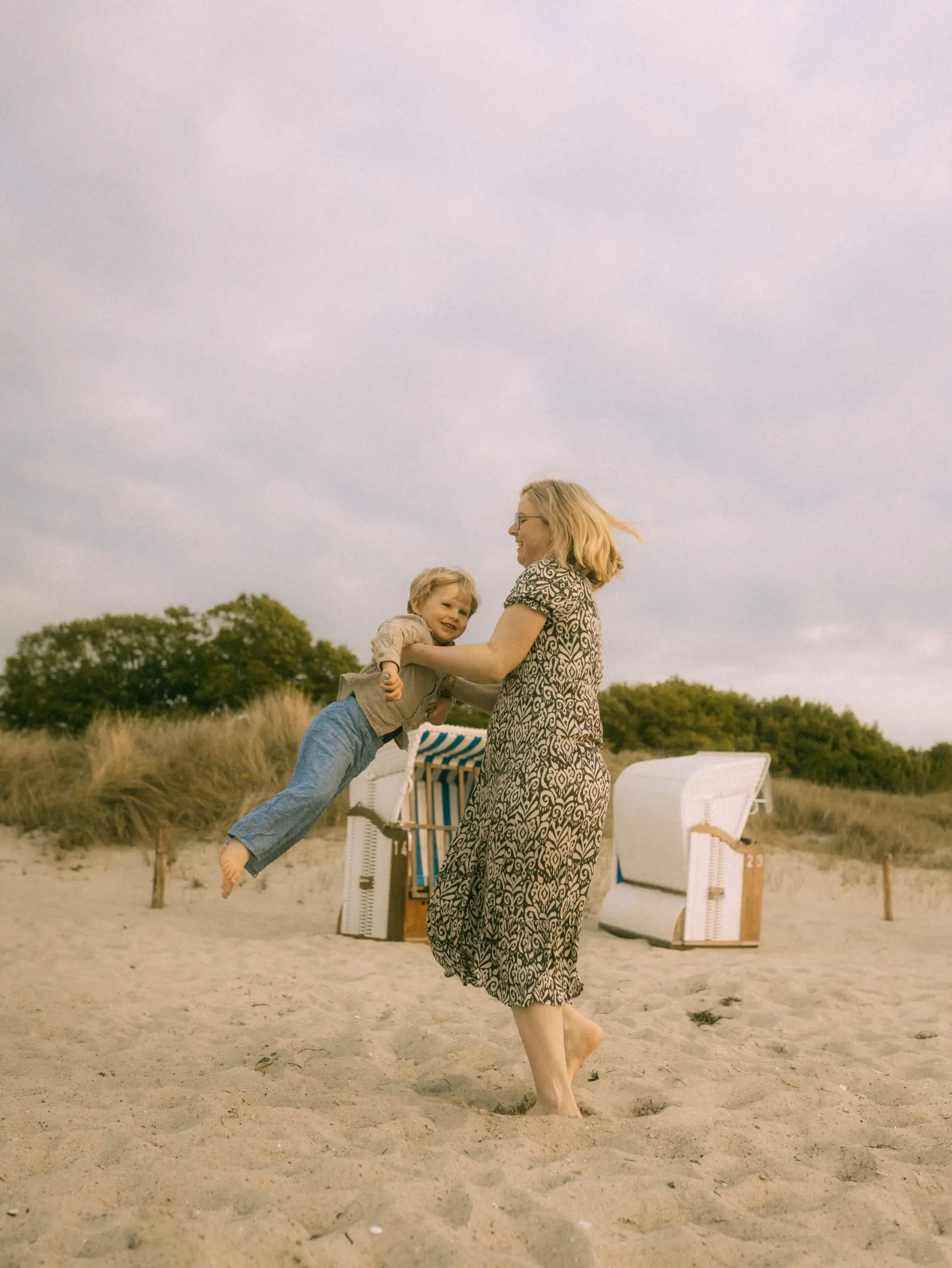Eine Mutter wirbelt lachend ihren kleinen Sohn am Strand herum, während sie gemeinsam einen fröhlichen Moment genießen. Im Hintergrund stehen typische Strandkörbe in den Dünen, was diese Familienfotos in Schwerin zu einer besonders schönen Erinnerung macht.