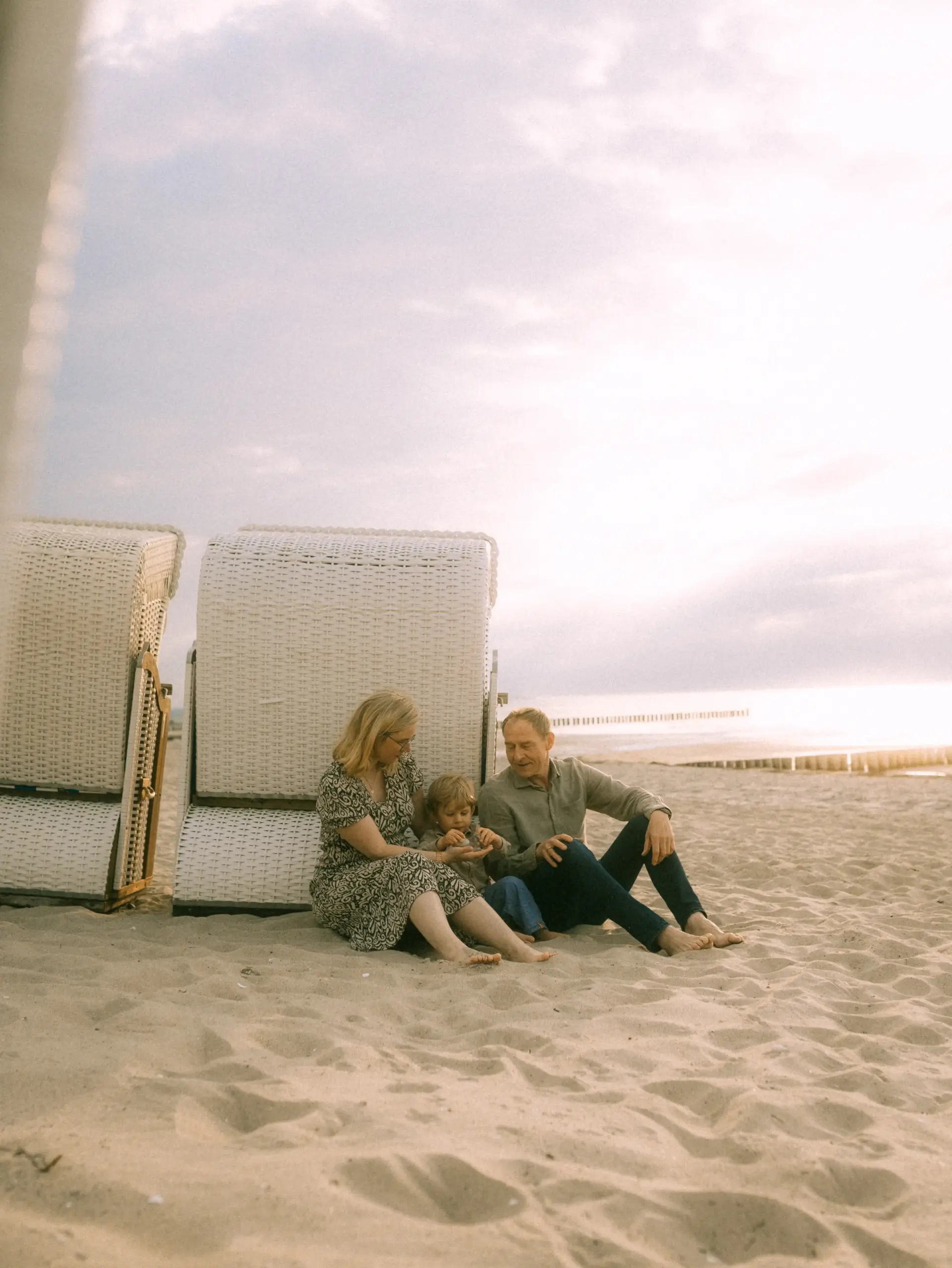 Eine junge Familie sitzt entspannt im Sand vor einem Strandkorb an der Ostsee. Die Eltern und ihr Kind genießen gemeinsam einen Moment am Meer. Authentische Familienfotos in Schwerin, die die Verbundenheit und das Glück im Urlaub einfangen.