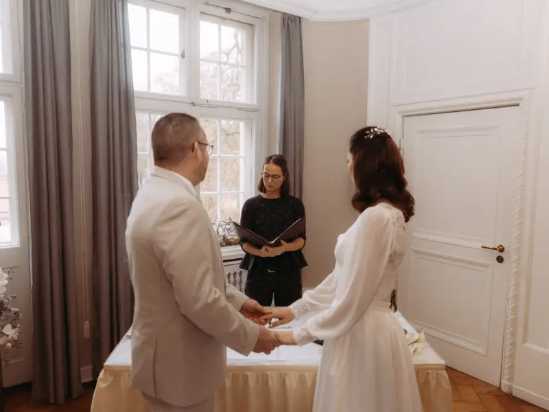 Ein Brautpaar hält sich während der standesamtlichen Trauung in einem hellen, eleganten Raum an den Händen, während die Standesbeamtin die Zeremonie leitet – festgehalten vom Hochzeitsfotograf in Schwerin und MV.