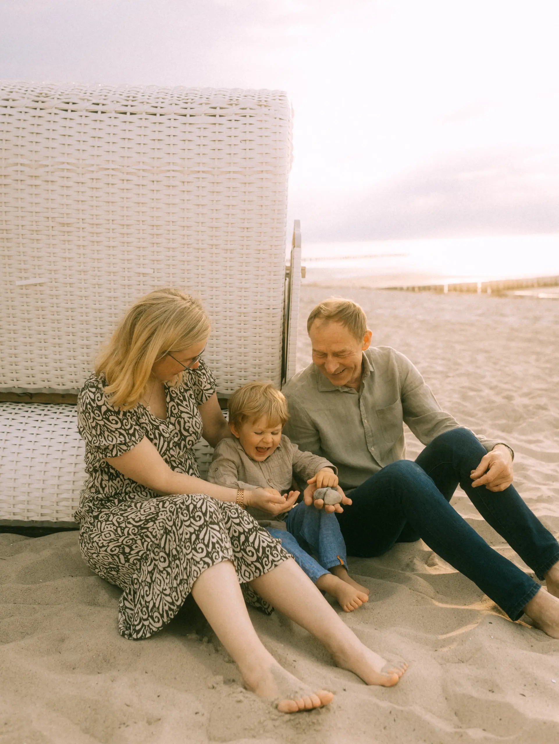 Ein glückliches Paar mit ihrem kleinen Sohn spielt gemeinsam im Sand vor einem Strandkorb. Die Familie genießt einen entspannten Moment am Meer im warmen Licht der Abendsonne. Authentische Familienfotos in Schwerin, die die Verbundenheit und Freude beim Spielen am Strand einfangen.