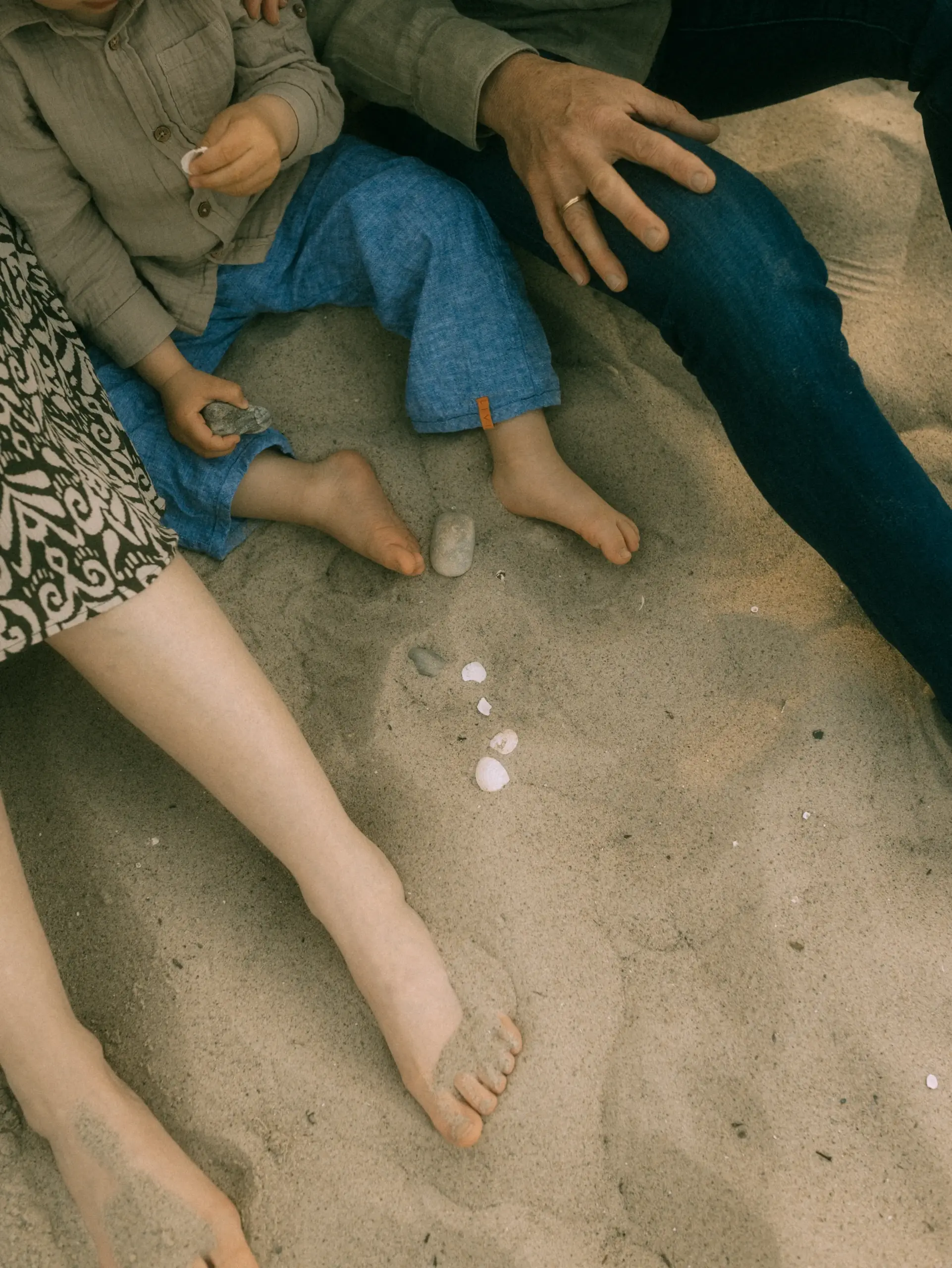Ein Familienfotograf in Schwerin hält einen intimen Moment am Strand fest: Ein Kind in Leinenkleidung und seine Eltern sitzen gemeinsam im Sand, spielen mit kleinen Steinen und Muscheln und genießen ihre Zeit bei einem entspannten Familienfotoshooting in Schwerin.