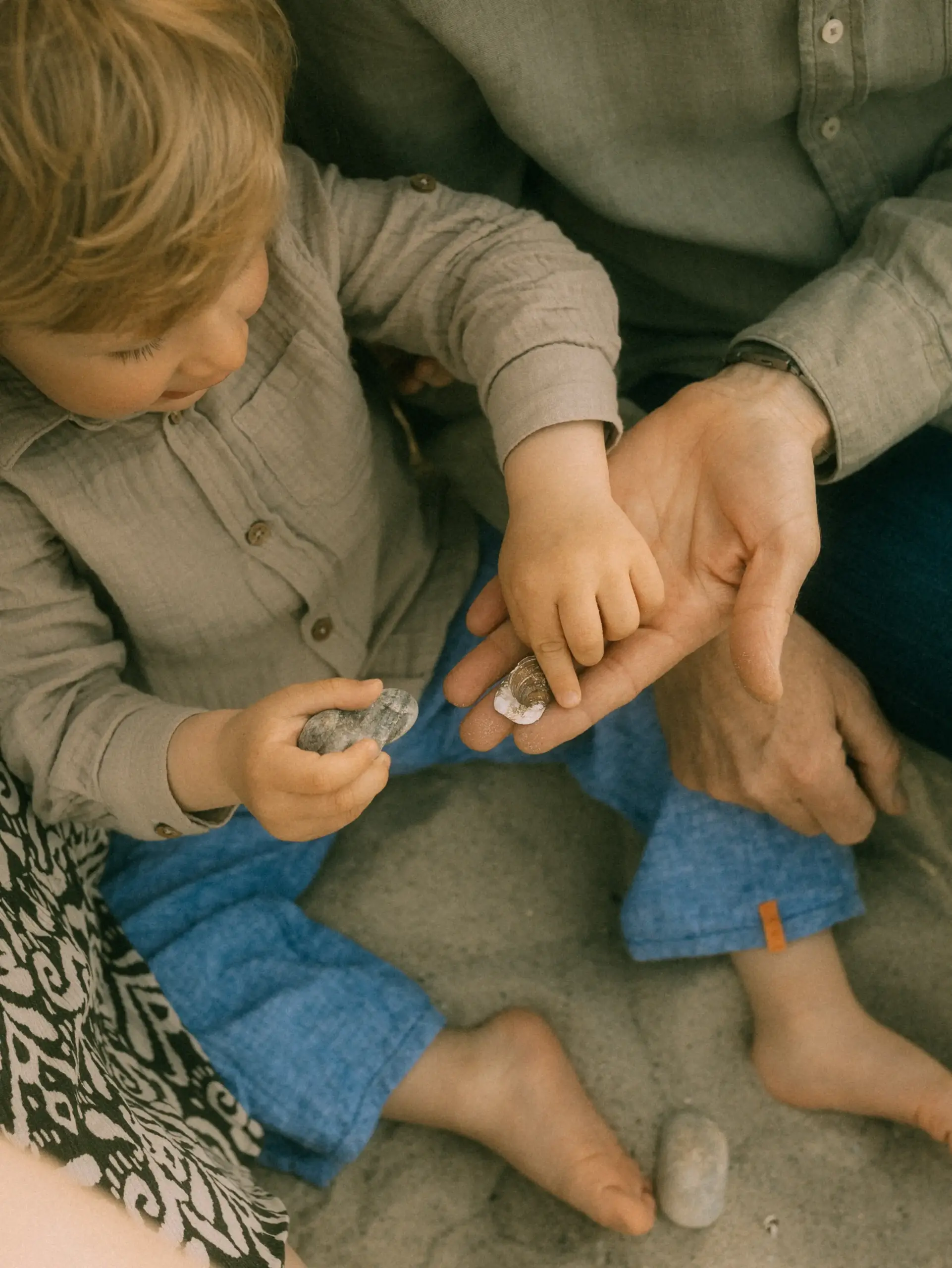 Ein Kleinkind in einem beigen Hemd sitzt am Strand und hält gemeinsam mit einem Erwachsenen kleine Muscheln und Steine in den Händen. Die Szene vermittelt eine liebevolle Verbindung und hält einen kostbaren Moment fest, wie ihn ein Familienfotograf in Schwerin einfängt.