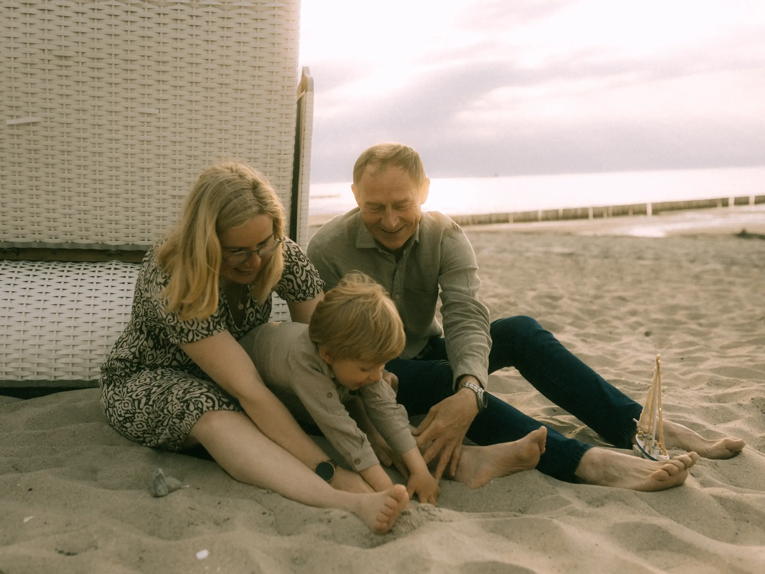 Ein glückliches Paar mit ihrem kleinen Sohn spielt gemeinsam im Sand vor einem Strandkorb an der Ostsee. Die Familie genießt einen entspannten Moment am Meer, während ein kleines Spielzeug-Segelschiff im Sand steht. Authentische Familienfotos in Schwerin.