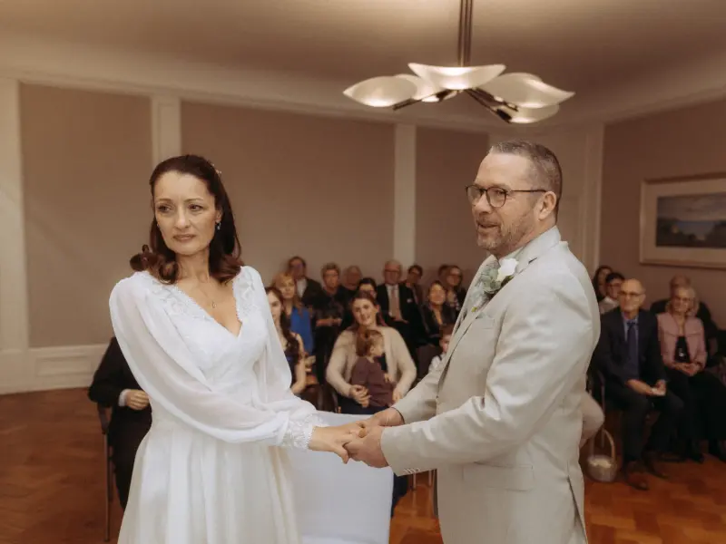 Ein Brautpaar hält sich während der Trauung in einem eleganten Saal an den Händen. Die Braut trägt ein weißes Kleid mit langen Ärmeln, der Bräutigam einen hellen Anzug. Im Hintergrund sitzen Hochzeitsgäste. Aufgenommen von einem Hochzeitsfotograf in Schwerin und MV.