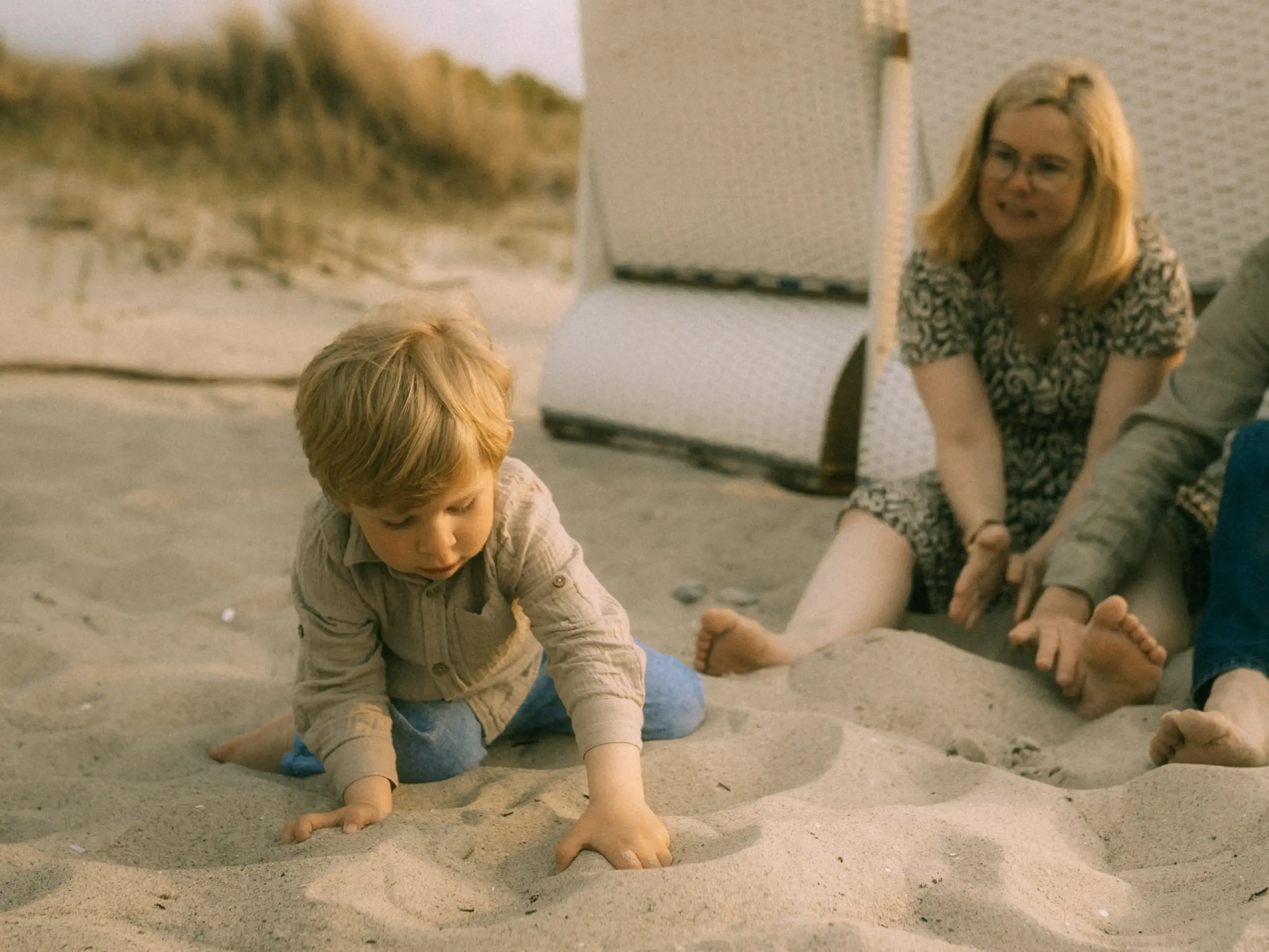 Ein kleines Kind spielt konzentriert im Sand am Strand, während die Mutter im Hintergrund zuschaut. Die entspannte Atmosphäre am Meer fängt einen wertvollen Familienmoment ein. Professionelle Aufnahmen wie diese bietet Ihr Familienfotograf in Schwerin.