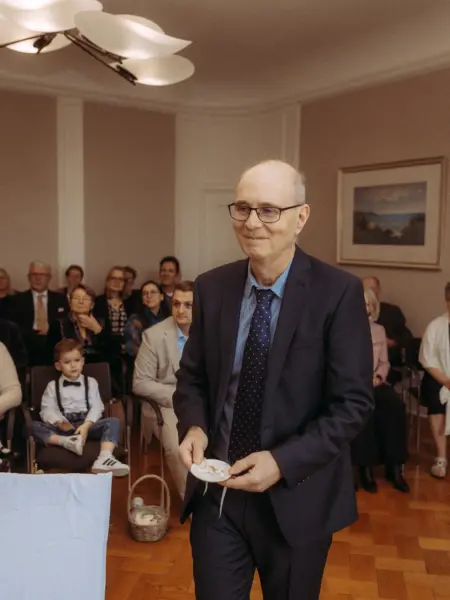 Ein lächelnder älterer Herr im dunkelblauen Anzug und mit Krawatte hält während einer Hochzeitszeremonie ein kleines weißes Tellerchen mit Eheringen in den Händen. Im Hintergrund sitzen Gäste in einem festlichen Saal. Authentische Aufnahme vom Hochzeitsfotograf in Schwerin und MV.