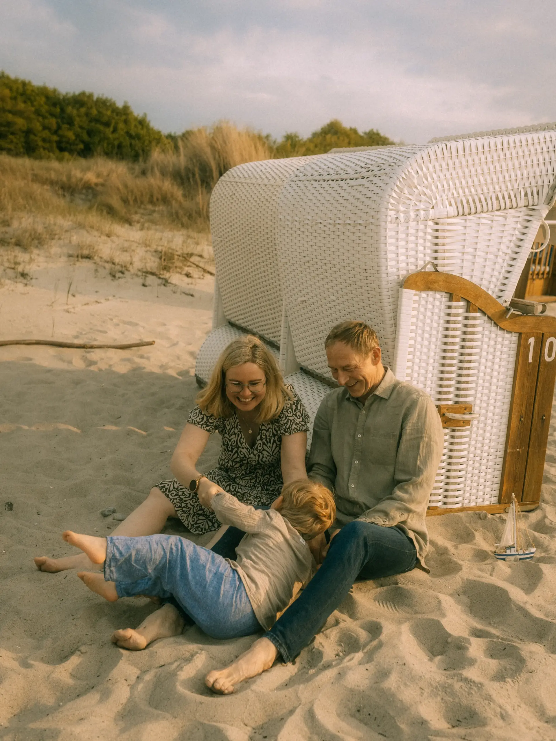 Glückliche Familie bei einem entspannten Strandtag vor einem weißen Strandkorb mit der Aufschrift „10“. Der Familienfotograf in Schwerin hat diesen natürlichen Moment eingefangen, während Eltern mit ihrem Kind im Sand spielen.