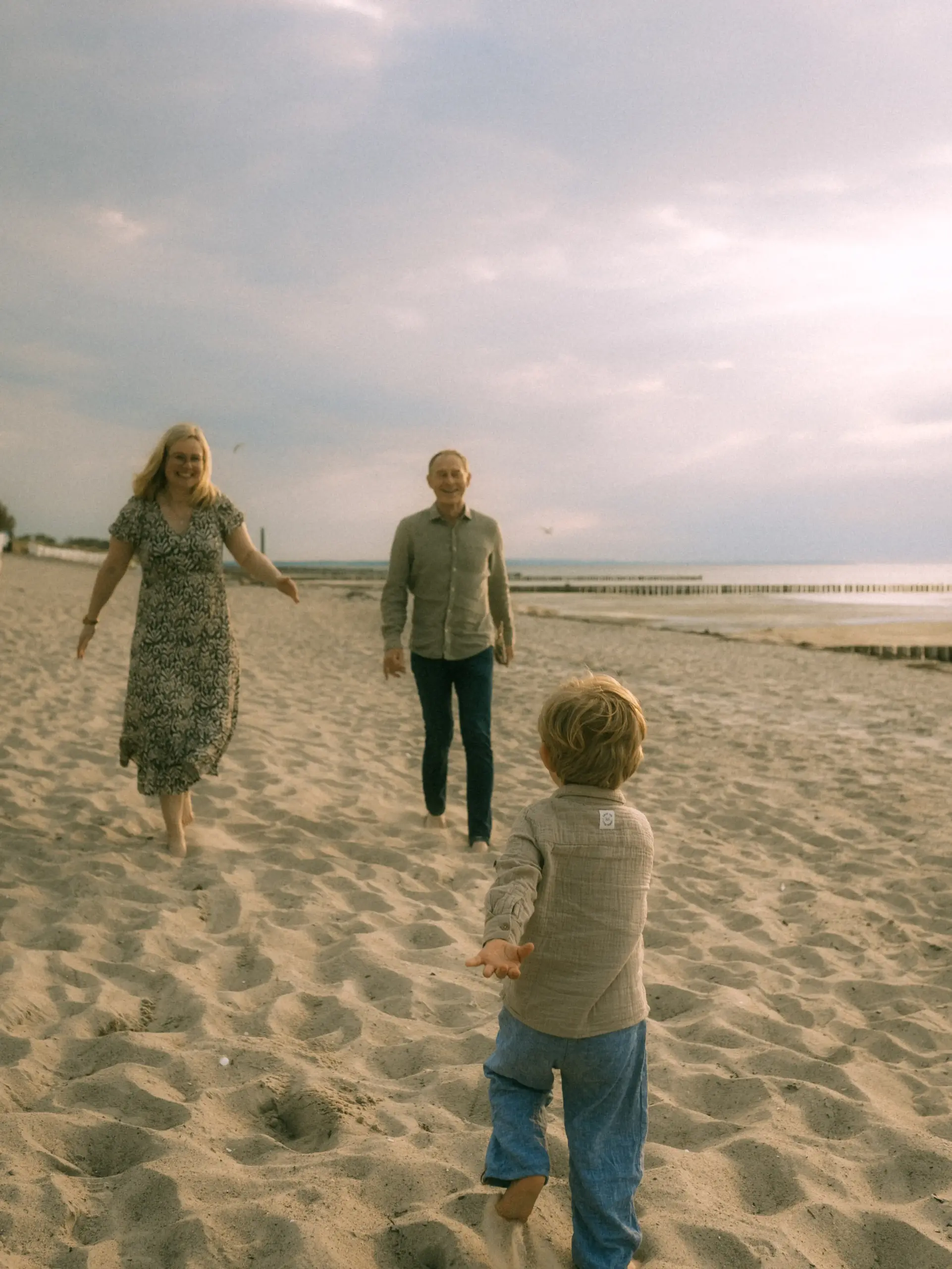 Ein kleiner Junge läuft freudig auf seine Eltern zu, die am Strand auf ihn warten. Die Familie genießt einen entspannten Tag am Meer in einer malerischen Küstenlandschaft. Authentische Familienfotos in Schwerin vom Profi eingefangen.