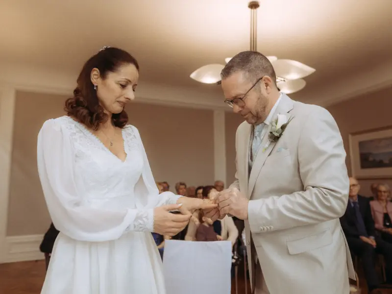 Eine Braut im weißen Spitzenkleid und ein Bräutigam im beigen Anzug tauschen während ihrer Trauung die Ringe. Der intime Moment wird von einem Hochzeitsfotograf in Schwerin und MV festgehalten, während Gäste im Hintergrund der Zeremonie in einem hellen Raum beiwohnen.