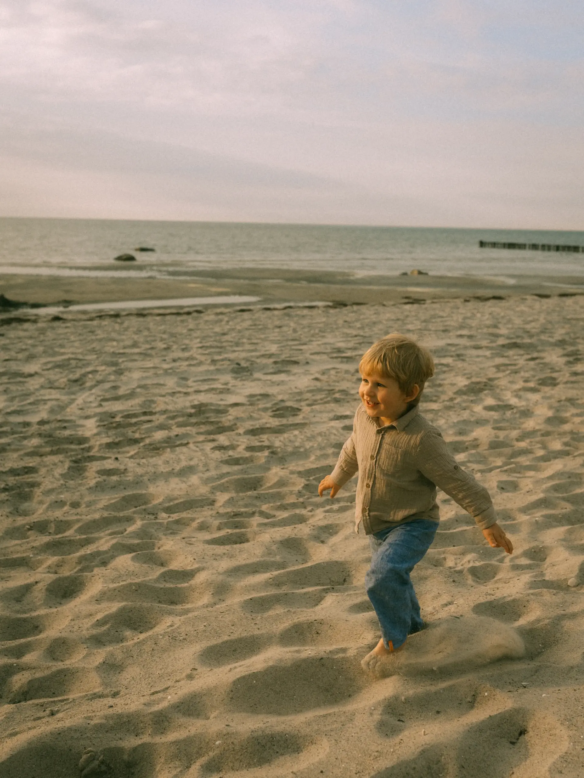 Ein kleiner Junge mit blonden Haaren läuft fröhlich barfuß über den Sandstrand am Meer. Er trägt ein beiges Hemd und eine blaue Hose. Die Szene fängt einen unbeschwerten Moment ein, ideal für Familienfotos in Schwerin.