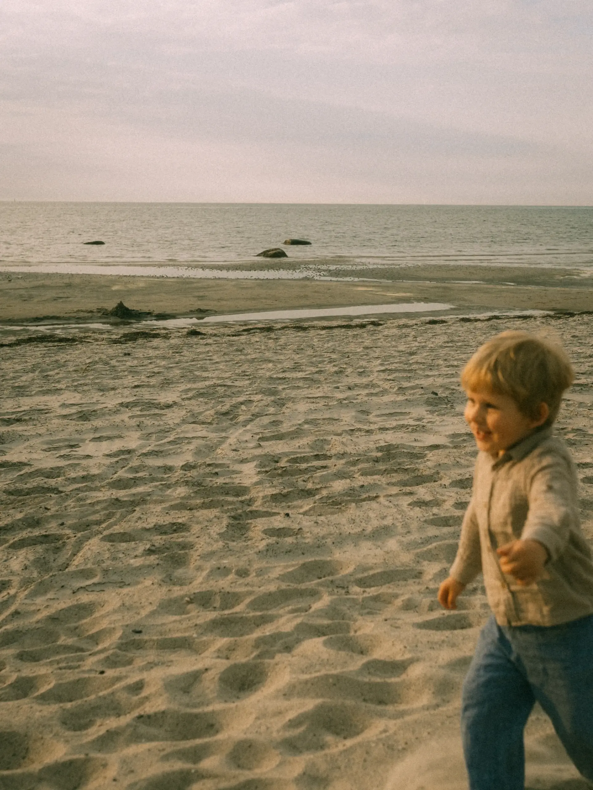 Ein kleiner Junge mit blonden Haaren rennt fröhlich über einen sandigen Strand am Meer, während er in ein beiges Hemd und eine blaue Hose gekleidet ist. Die Aufnahme fängt einen natürlichen Moment ein, wie ihn ein Fotograf für Familien in Schwerin bei einem Strandspaziergang festhalten würde.