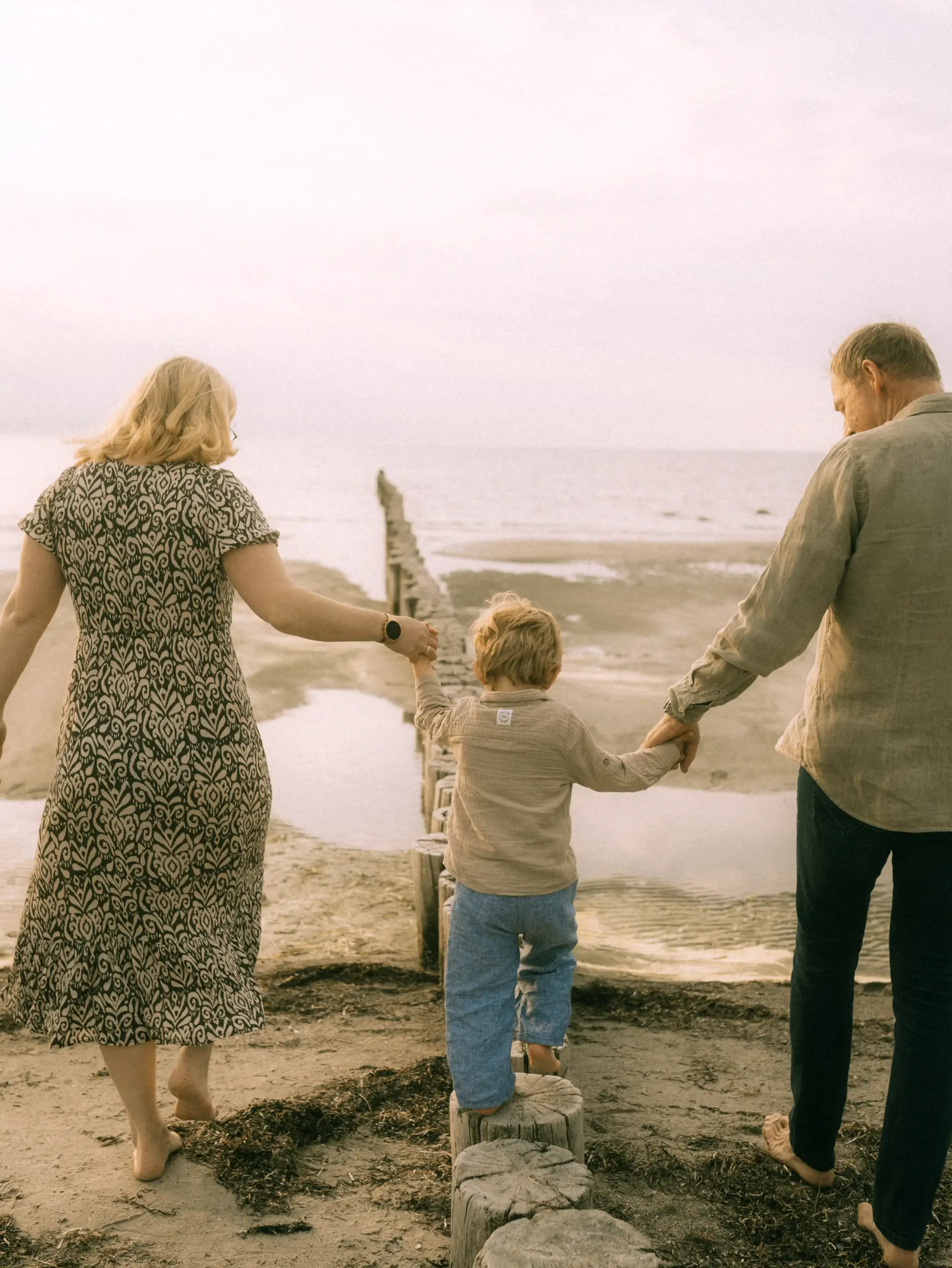 Ein Familienfotograf in Schwerin hält diesen Moment fest: Eine Mutter, ein Vater und ihr kleiner Sohn spazieren barfuß am Strand entlang und halten sich an den Händen, während sie über die hölzernen Wellenbrecher balancieren. Die Szene strahlt eine warme, friedliche Atmosphäre am Meer aus.