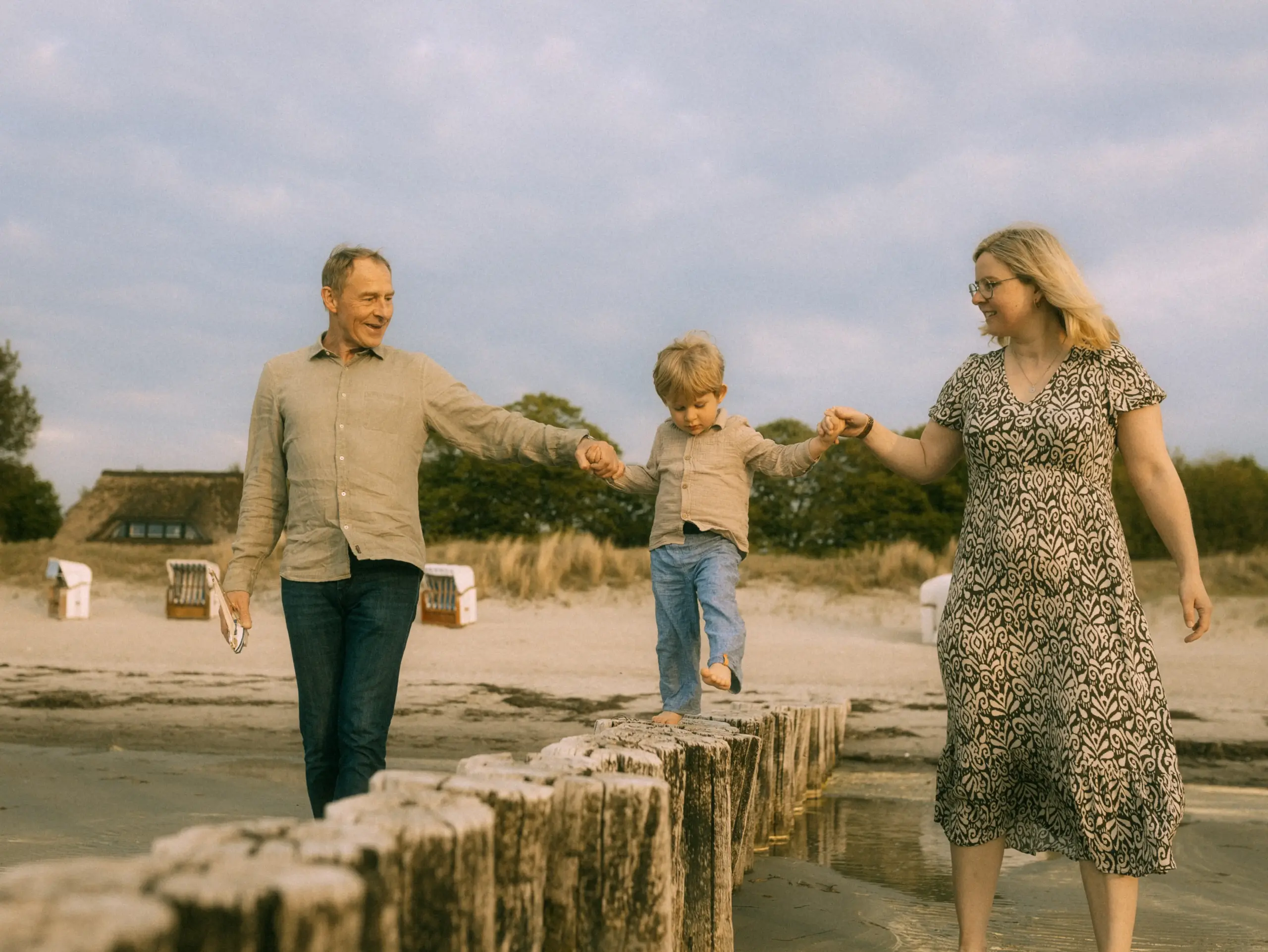 Ein Familienfotograf in Schwerin hält diesen besonderen Moment fest: Ein kleines Kind balanciert barfuß auf einer Reihe von Holzpfählen am Strand, während es von seinen Eltern an den Händen gehalten wird, umgeben von einer friedlichen Küstenlandschaft.
