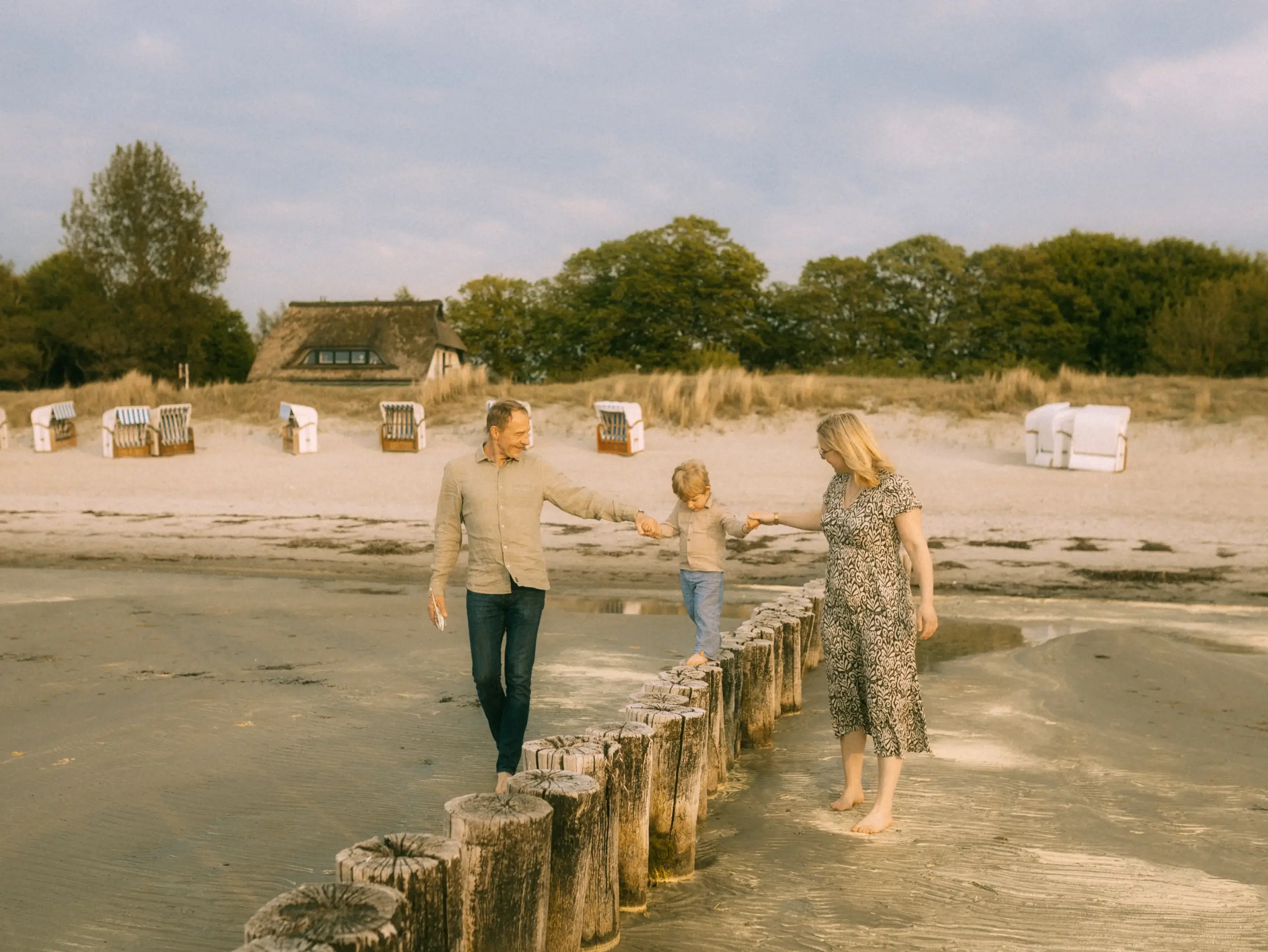 Eine junge Familie bei einem Strandspaziergang: Eltern halten ihr kleines Kind an den Händen, während es balancierend über eine Reihe von Holzpfählen im Sand läuft. Im Hintergrund stehen typische Strandkörbe vor einer Dünenlandschaft. Familienfotograf in Schwerin.