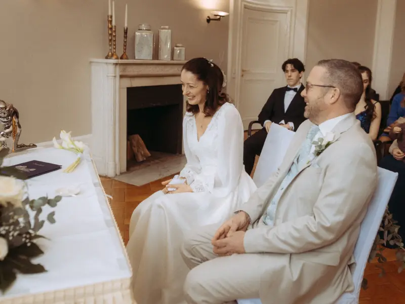 Ein glückliches Brautpaar sitzt während der standesamtlichen Trauung an einem festlich gedeckten Tisch. Die Braut im weißen Kleid und der Bräutigam im hellen Anzug lächeln sich an. Professionelle Begleitung durch einen Hochzeitsfotograf in Schwerin und MV in einem eleganten historischen Raum.