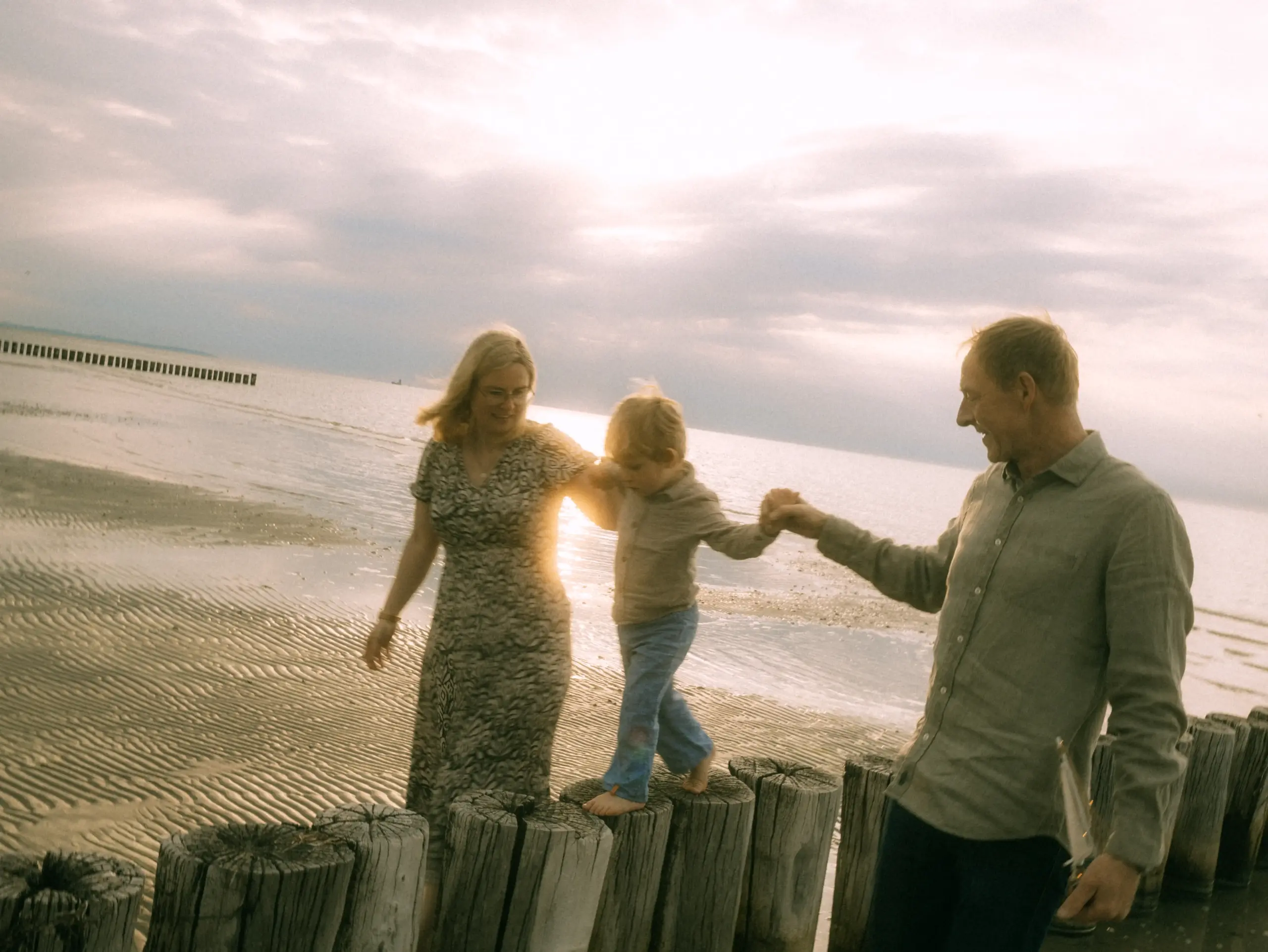 Eine glückliche Familie balanciert bei Sonnenuntergang gemeinsam über eine Reihe von Holzpfählen am Strand. Als Familienfotograf in Schwerin fange ich diesen emotionalen Moment fest, der die Verbundenheit zwischen Eltern und Kind in einer malerischen Küstenlandschaft zeigt.