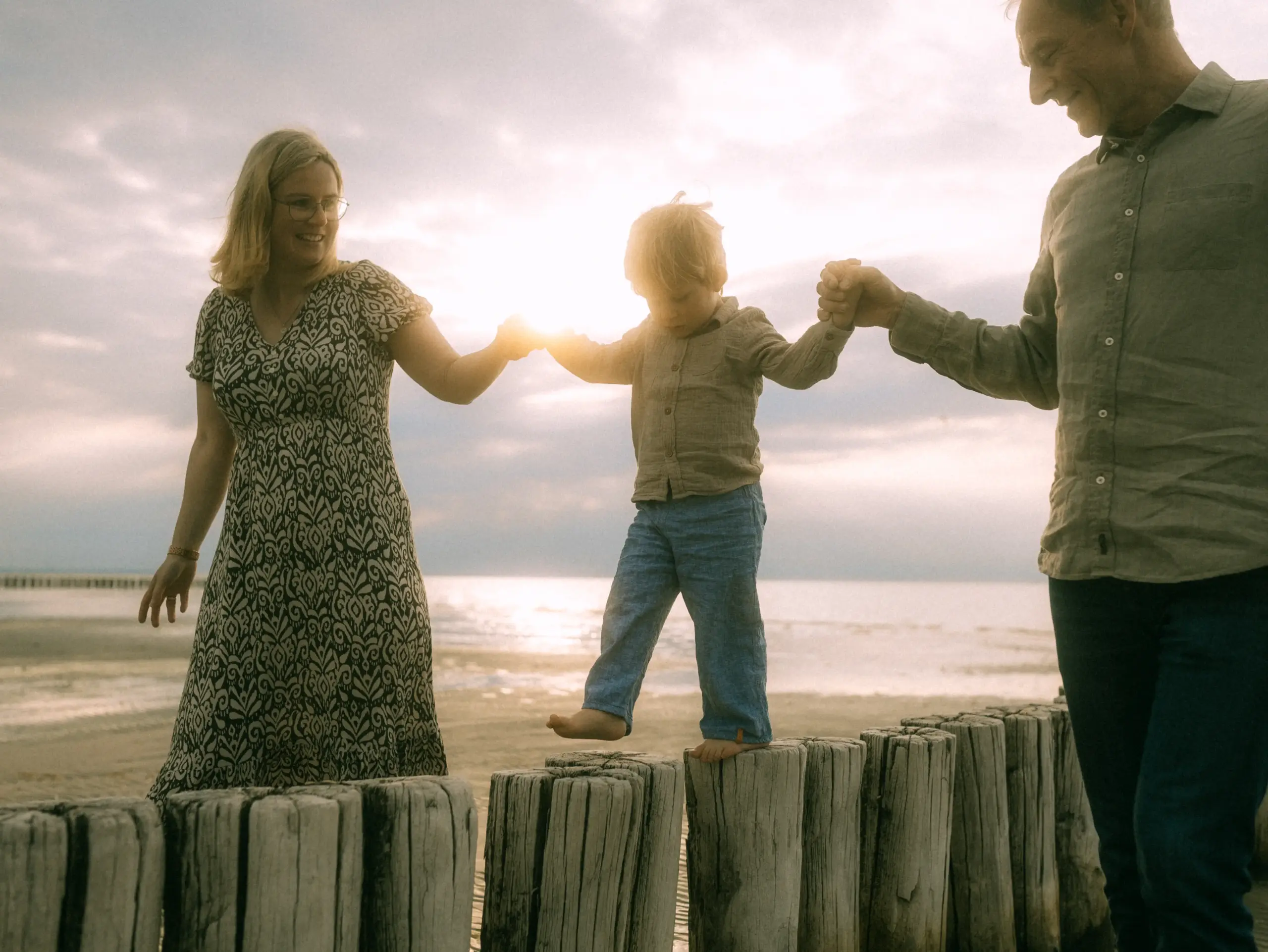 Ein kleiner Junge balanciert auf einer Reihe von Holzpfählen am Strand, während seine Eltern ihn an beiden Händen halten. Die Familie genießt einen sonnigen Moment am Meer, eingefangen von einem Fotograf für Familien in Schwerin in einer stimmungsvollen Gegenlichtaufnahme.