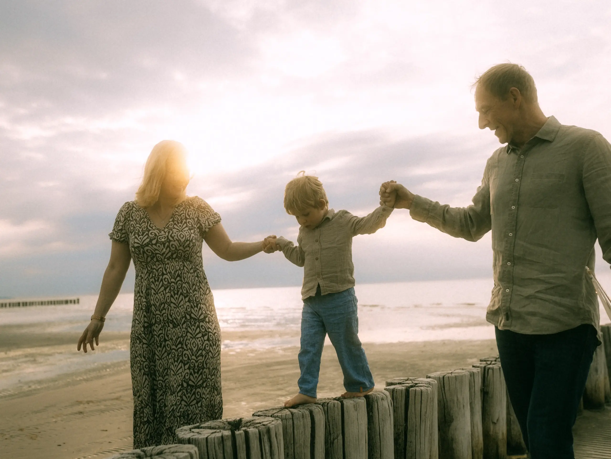 Ein kleiner Junge balanciert barfuß auf einer Holzpfahlreihe am Strand, während er von seinen Großeltern an den Händen gehalten wird. Die warme Abendsonne taucht die Szene in goldenes Licht. Ein herzlicher Moment, festgehalten von einem Fotograf für Familien in Schwerin.