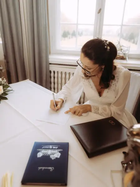 Eine Braut in einem eleganten weißen Spitzenkleid unterschreibt konzentriert die Heiratsurkunde während ihrer standesamtlichen Trauung. Auf dem Tisch liegen eine blaue Hochzeitsmappe mit der Aufschrift 