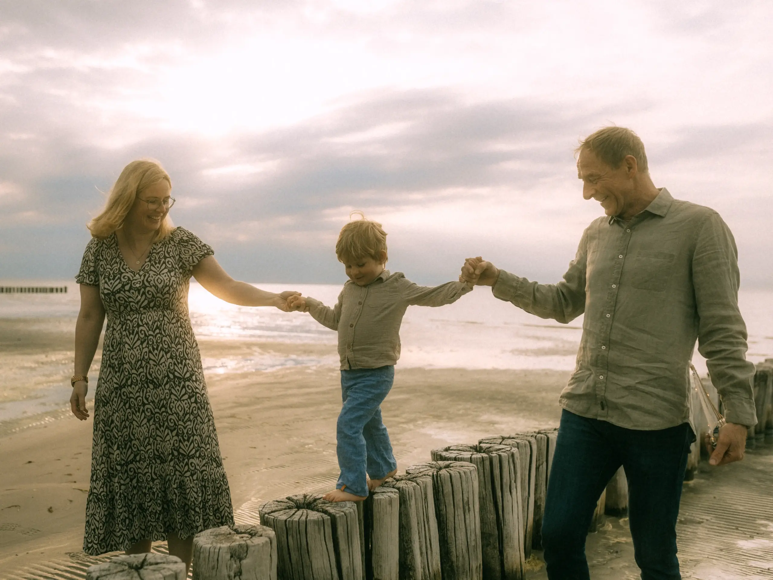 Eine blonde Frau und ein Mann halten einen kleinen Jungen an den Händen, während er barfuß über eine Reihe von Holzpfählen am Strand balanciert. Die Familie genießt einen sonnigen Moment am Meer, eingefangen von einem professionellen Familienfotograf in Schwerin.