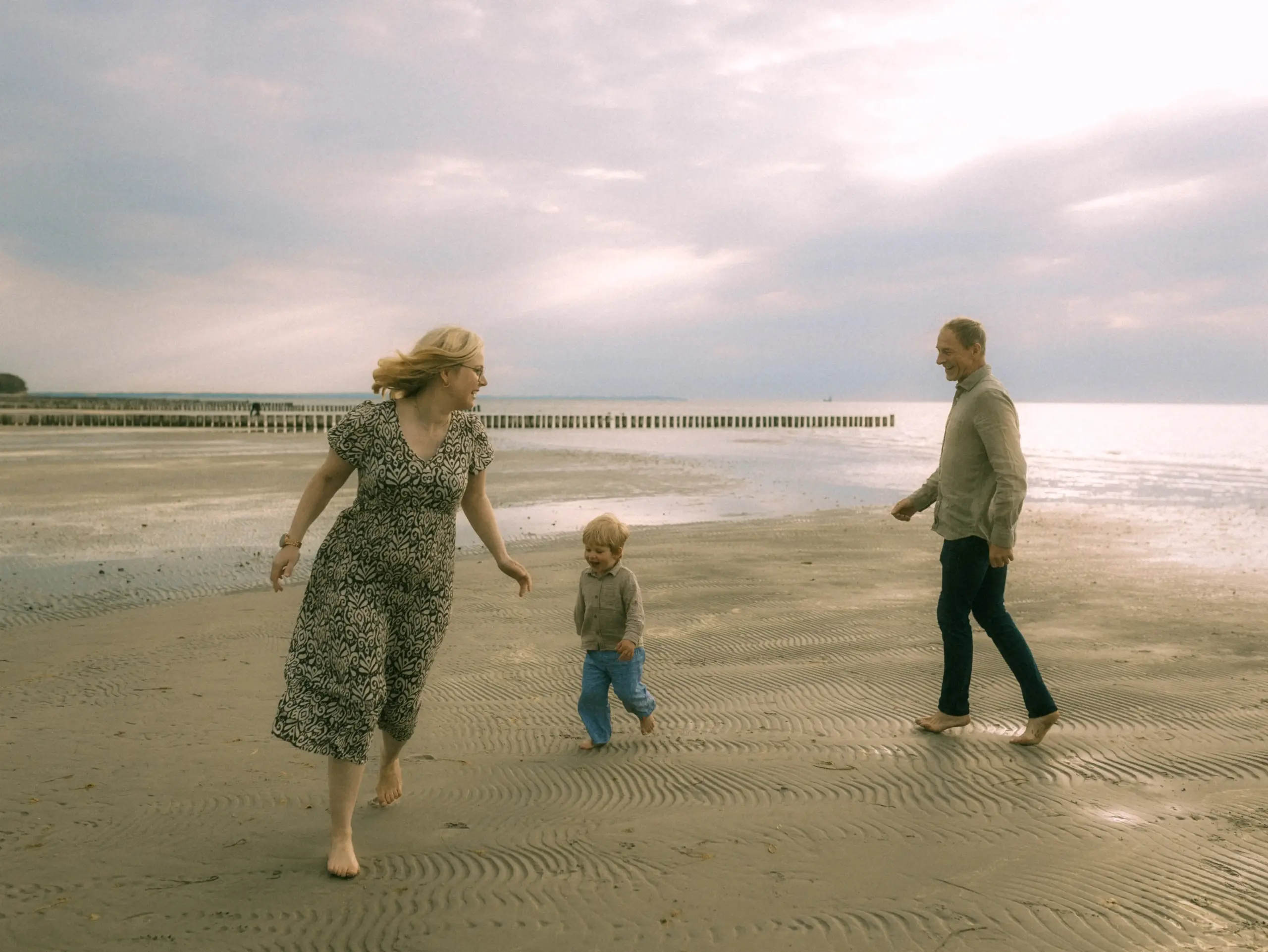 Eine glückliche Familie läuft barfuß am Strand entlang. Mutter, Vater und Kind lachen gemeinsam beim Spielen im Sand unter einem bewölkten Himmel. Authentische Familienfotos in Schwerin, die eine fröhliche Stimmung und die Verbundenheit bei einem Spaziergang am Meer einfangen.