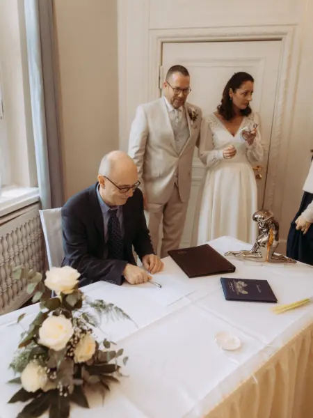 Ein Standesbeamter unterschreibt die Heiratsurkunde, während das Brautpaar aufmerksam zuschaut. Auf dem Tisch steht eine silberne Skulptur und ein blaues Dokumentenheft mit der Aufschrift „Standesamt“. Authentische Momente festgehalten vom Hochzeitsfotograf in Schwerin und MV.