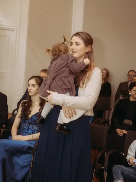 Eine junge Frau hält liebevoll ein Kleinkind auf dem Arm, während sie bei einer festlichen Veranstaltung in einem Saal steht. Im Hintergrund sitzen weitere Gäste in eleganter Kleidung. Professionelle Fotografie für Familien in Schwerin, die emotionale Momente festhält.