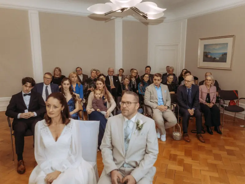 Brautpaar bei der standesamtlichen Trauung im eleganten Raum mit Gästen im Hintergrund, fotografiert von einem Hochzeitsfotograf in Schwerin und MV. Die Braut trägt ein weißes Spitzenkleid, der Bräutigam einen hellen Anzug, während die Hochzeitsgesellschaft aufmerksam zuschaut.