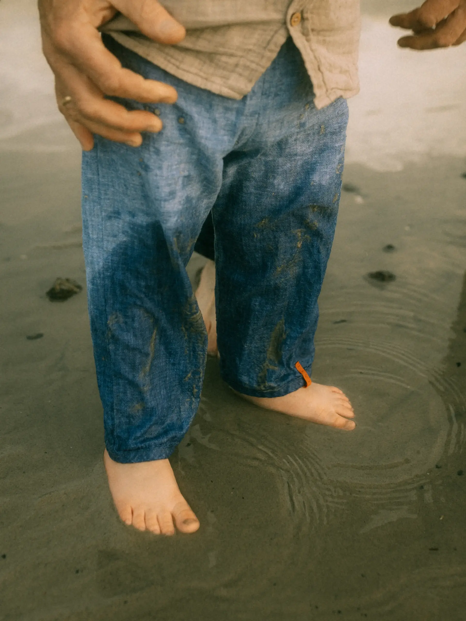 Ein Paar steht barfuß im Wasser, die Füße eines Kindes mit blauen Jeans und einer orangefarbenen Schnur am Knöchel sind sichtbar.