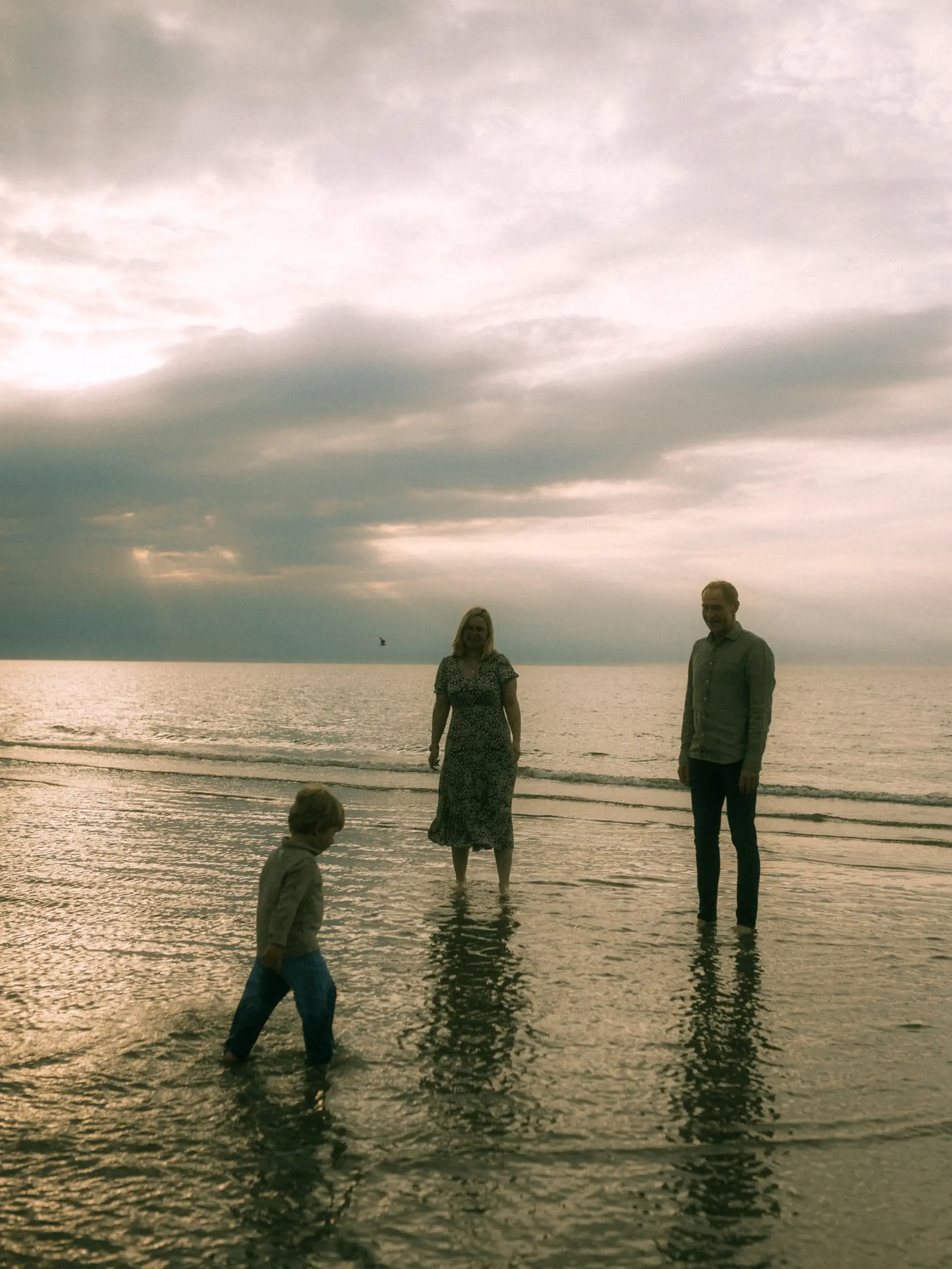 Eine Familie steht bei Sonnenuntergang im flachen Wasser am Strand. Ein kleiner Junge spielt im Vordergrund, während die Eltern im Hintergrund stehen. Die stimmungsvolle Atmosphäre am Meer unter einem bewölkten Himmel unterstreicht diese Familienfotos in Schwerin.