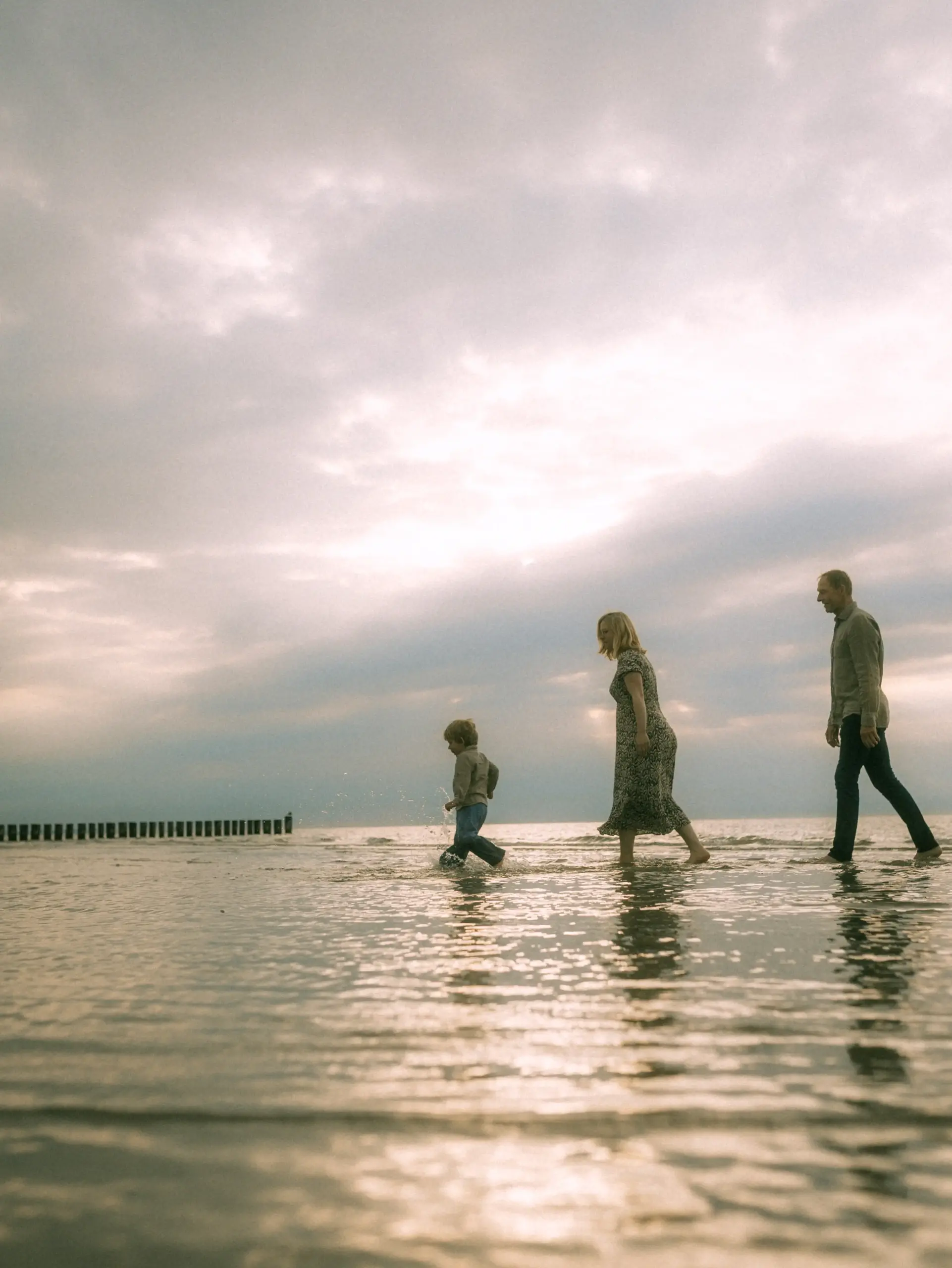 Eine junge Familie genießt einen entspannten Spaziergang am flachen Meeresufer während eines bewölkten Sonnenuntergangs. Das Kind spielt im seichten Wasser, während die Eltern daneben hergehen. Authentische Familienfotos in Schwerin, die eine emotionale Verbindung in der Natur zeigen.