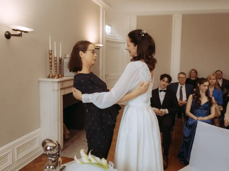 Eine Braut im weißen Brautkleid umarmt herzlich eine Frau in einem dunkelblauen gepunkteten Outfit während einer intimen Hochzeitszeremonie. Im Hintergrund sitzen Gäste in einem eleganten Raum. Hochzeitsfotograf in Schwerin und MV.