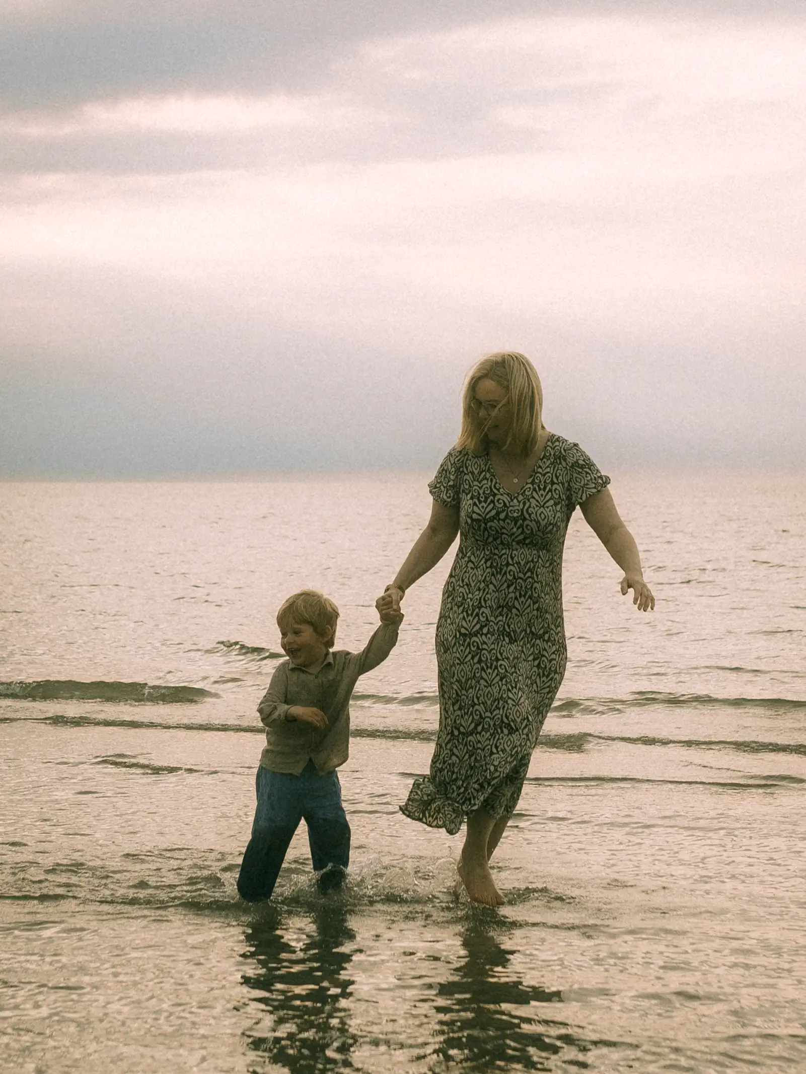 Eine Mutter und ihr kleiner Sohn halten Händchen und laufen fröhlich durch das flache Wasser am Strand bei bewölktem Himmel. Das emotionale Familienfoto fängt einen glücklichen Moment am Meer ein. Familienfotograf in Schwerin.