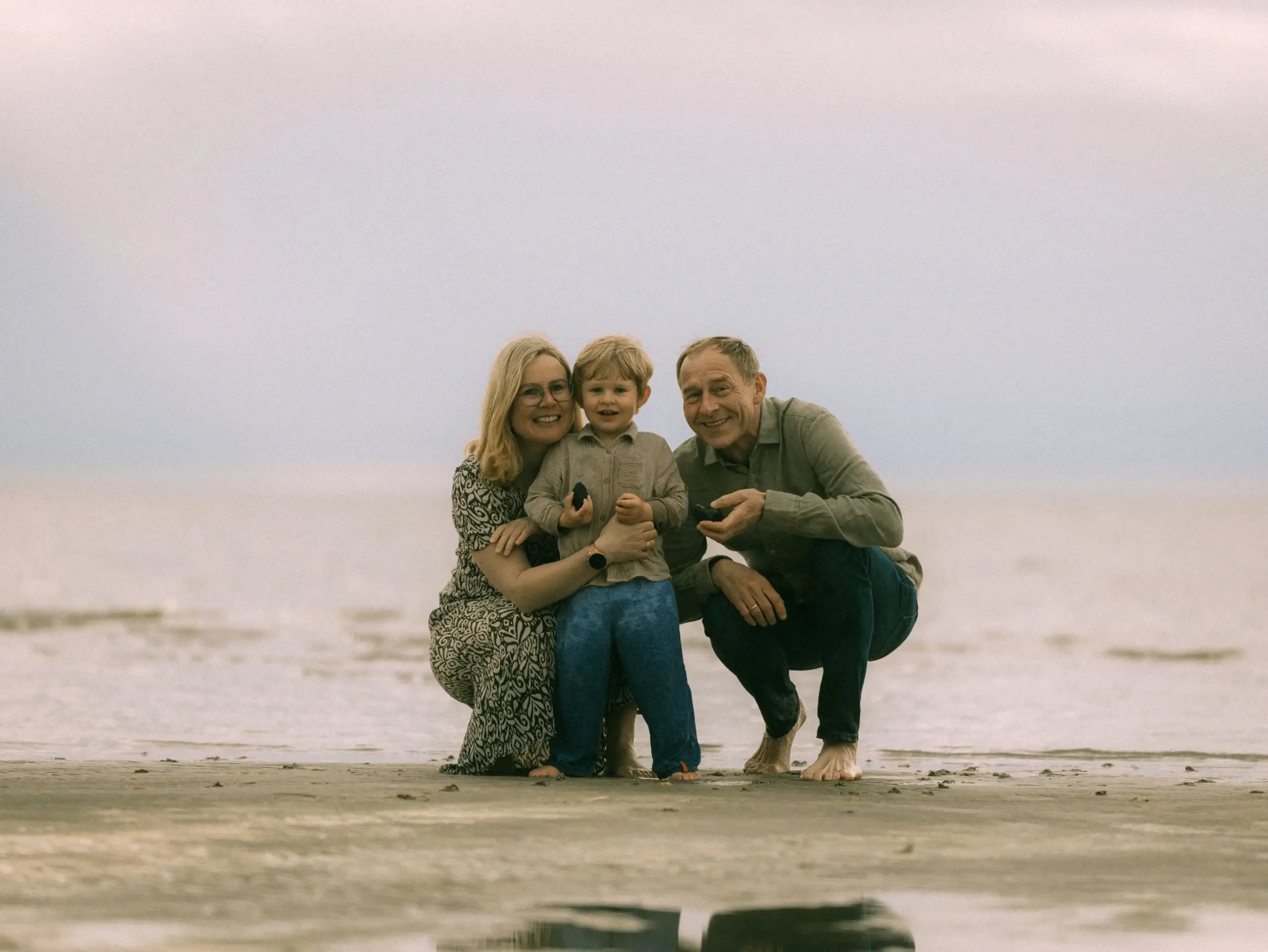 Eine glückliche Familie, bestehend aus Mutter, Vater und ihrem kleinen Sohn, kniet gemeinsam am flachen Sandstrand bei bewölktem Himmel. Die drei lächeln in die Kamera, während sie entspannte Zeit am Meer verbringen – ein schönes Motiv für Familienfotos in Schwerin.