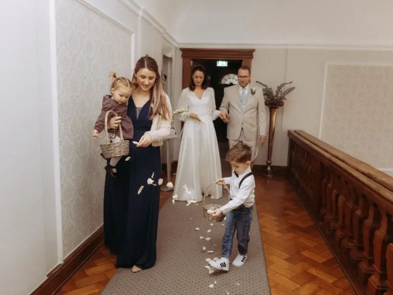 Ein Blumenmädchen und ein kleiner Junge streuen Blütenblätter auf den Gang, während das Brautpaar im Hintergrund folgt. Eine elegante Szene festgehalten von einem Hochzeitsfotograf in Schwerin und MV in einem historischen Gebäude mit Parkettboden und hellem Flur.