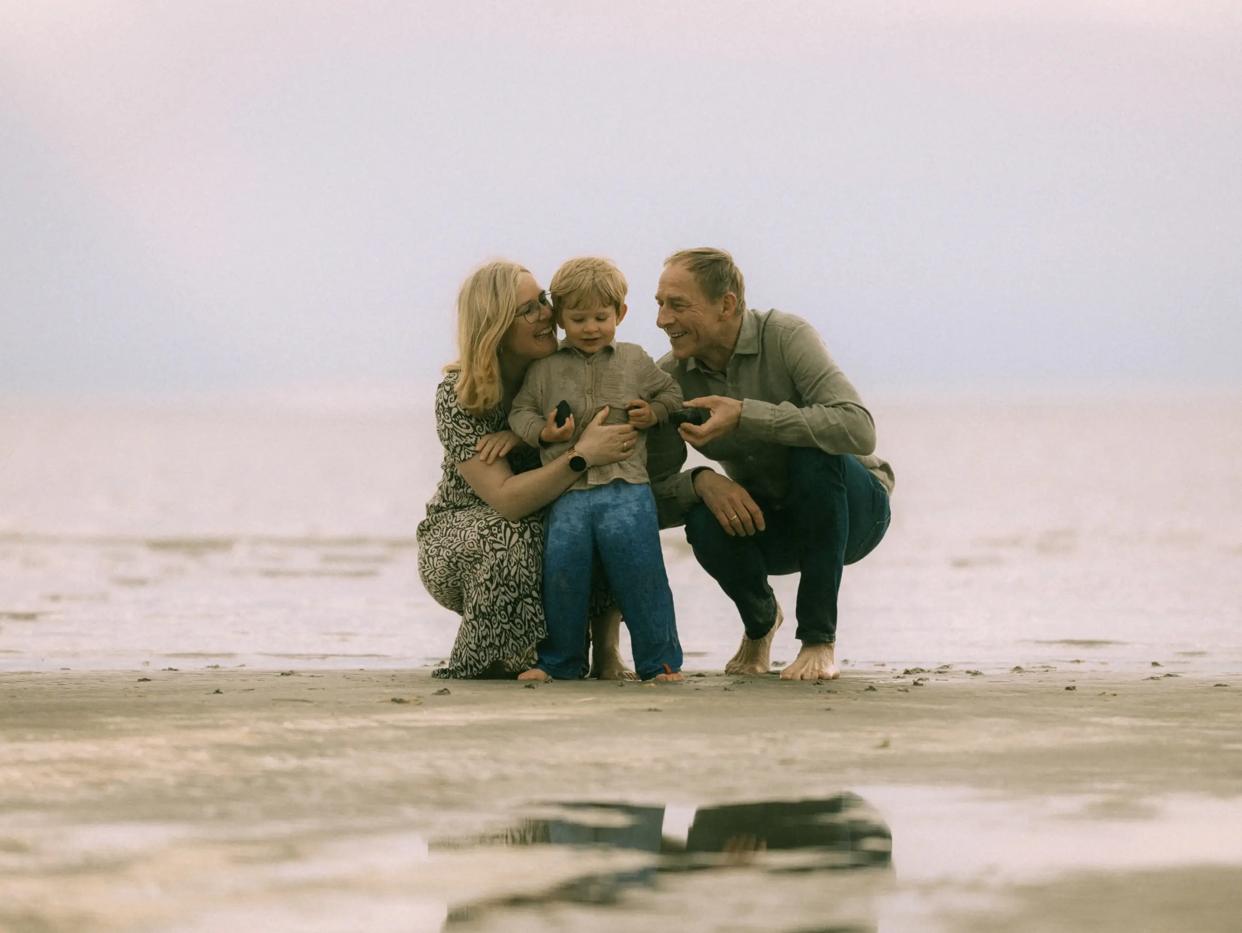 Eine glückliche Familie, bestehend aus Eltern und ihrem kleinen Sohn, verbringt einen entspannten Tag am Strand. Die drei knien auf dem nassen Sand, während sie gemeinsam spielen. Authentische Familienfotos in Schwerin, die liebevolle Momente am Wasser einfangen.