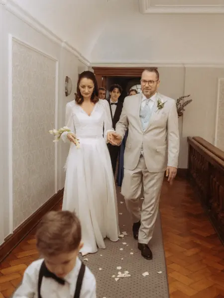 Eine Braut im weißen Kleid und ein Bräutigam im hellen Anzug schreiten Hand in Hand durch einen Flur, während ein kleiner Junge im Vordergrund den Weg bereitet. Diese emotionale Aufnahme wurde von einem Hochzeitsfotograf in Schwerin und MV festgehalten.