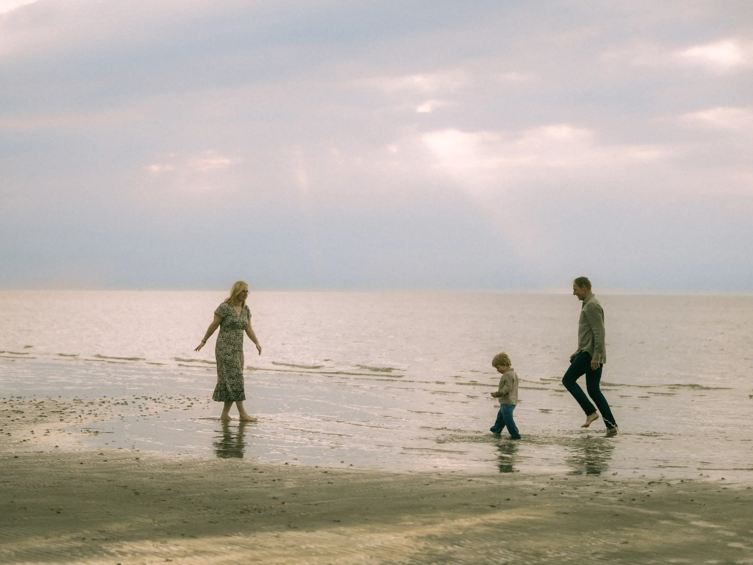 Eine junge Familie genießt einen entspannten Moment am Strand bei Sonnenuntergang. Die Mutter in einem gemusterten Kleid, der Vater und ihr kleiner Sohn laufen barfuß durch das flache Wasser. Ein stimmungsvolles Familienfoto, festgehalten von einem Fotograf für Familien in Schwerin.