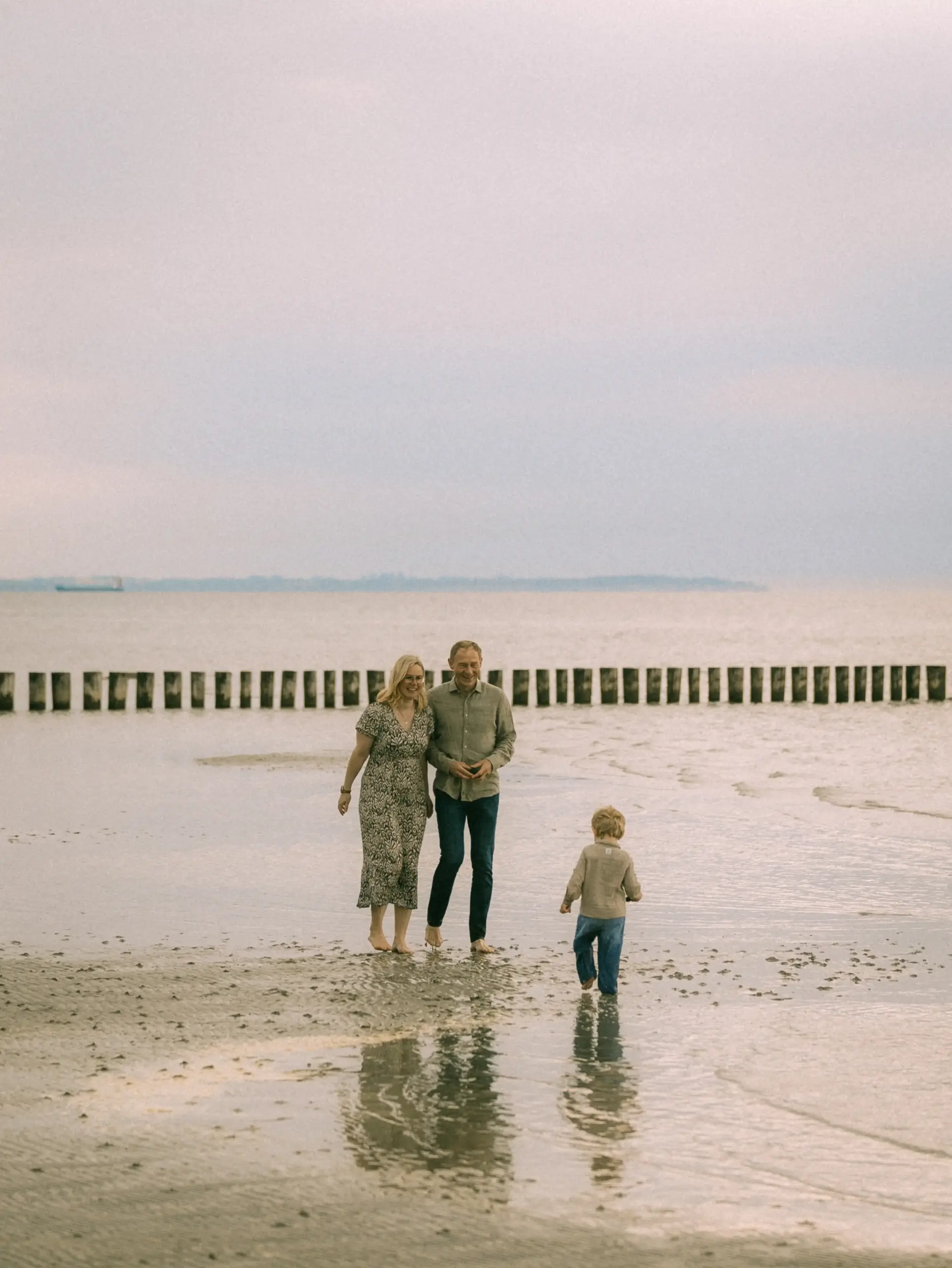 Eine junge Familie spaziert bei Ebbe barfuß am flachen Sandstrand entlang. Während die Eltern Hand in Hand gehen, läuft ihr kleiner Sohn voraus in Richtung Wasser. Im Hintergrund erstreckt sich eine lange Buhnenreihe im Meer. Authentische Familienfotos in Schwerin an der Küste.