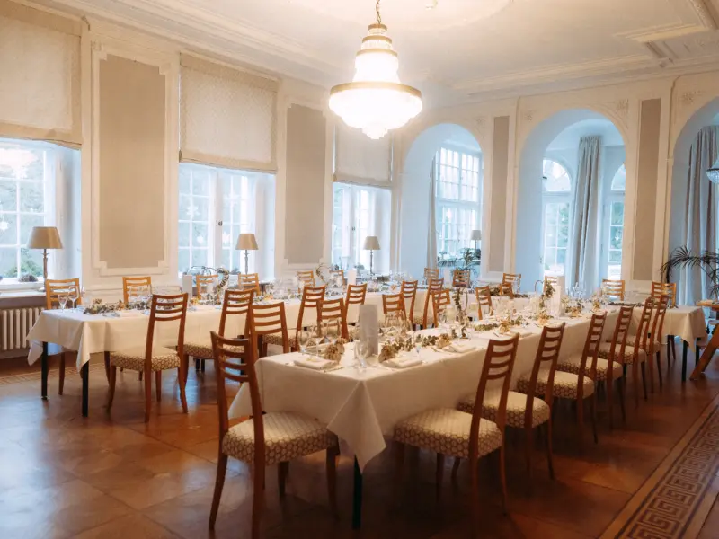 Ein eleganter, festlich gedeckter Festsaal für eine Hochzeitsfeier mit langen Tafeln, weißen Tischdecken, hellen Holzstühlen und einem prunkvollen Kronleuchter. Das Ambiente wirkt hell und einladend, ideal für einen Hochzeitsfotograf in Schwerin und MV.