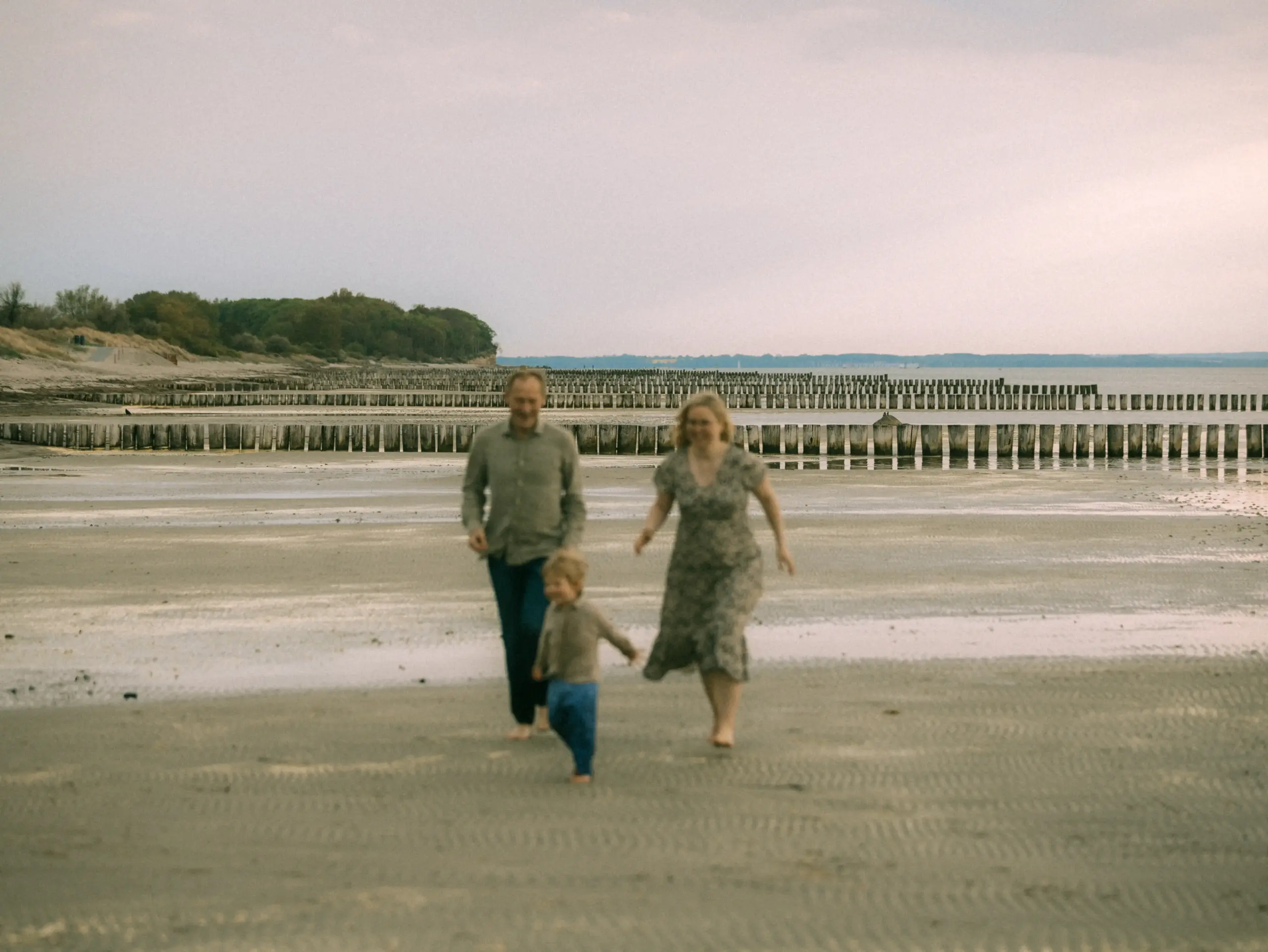 Eine glückliche Familie rennt barfuß und lachend über den flachen Sandstrand an der Ostsee. Die Eltern und ihr kleines Kind genießen einen entspannten Tag am Meer vor der Kulisse langer Buhnenreihen. Authentische Familienfotos in Schwerin und Umgebung.