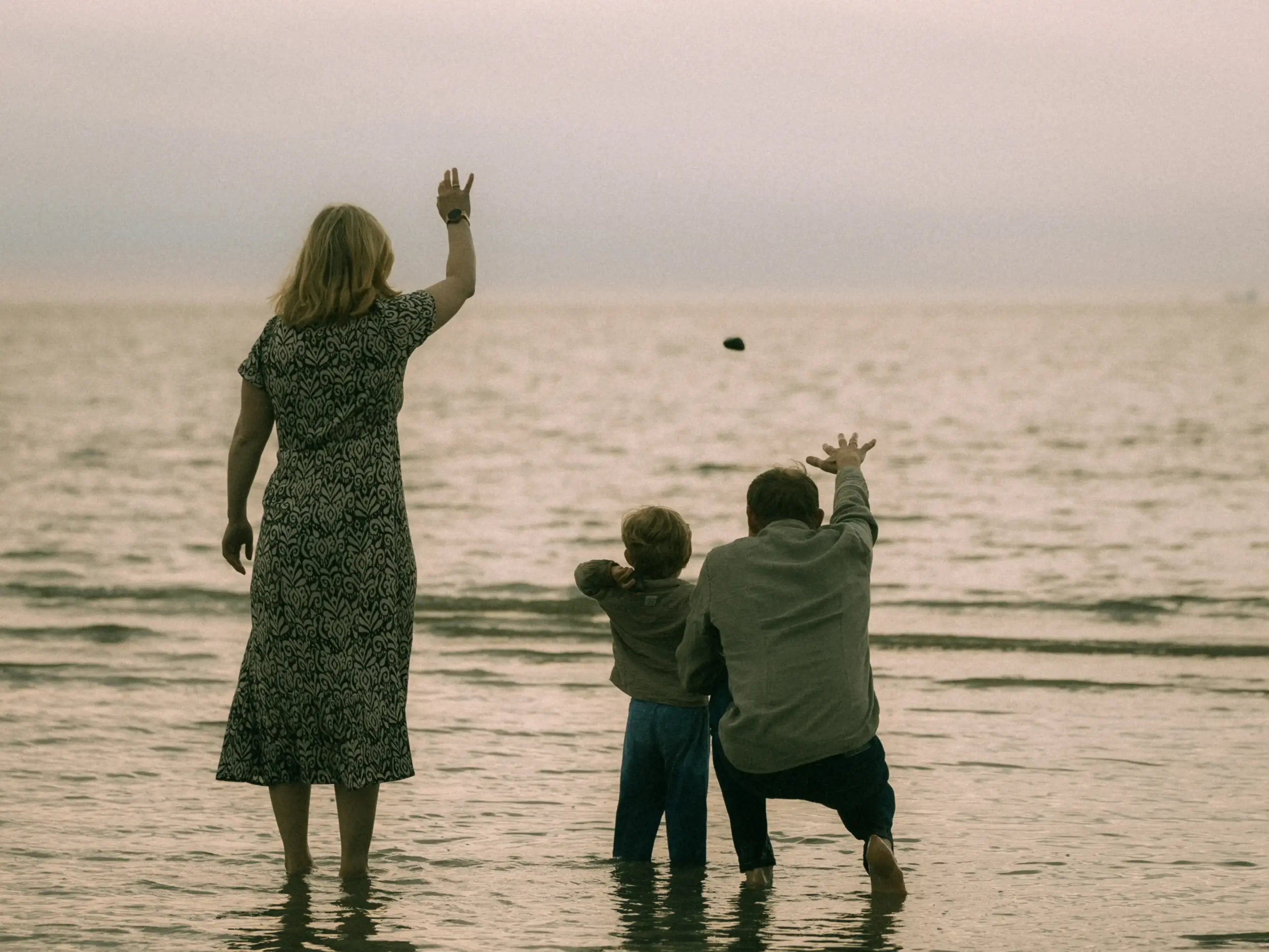 Eine Familie steht bei bedecktem Himmel im flachen Wasser am Strand und wirft gemeinsam Steine in das Meer. Die Szene fängt einen innigen Moment ein, der perfekt als Familienfotos in Schwerin festgehalten wurde.