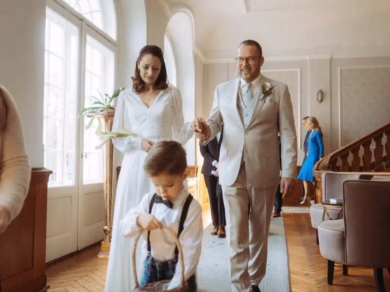 Ein Brautpaar schreitet Hand in Hand durch einen eleganten Saal, begleitet von einem kleinen Blumenjungen im Vordergrund. Die festliche Atmosphäre wird durch das klassische Hochzeitsoutfit unterstrichen. Professionelle Aufnahme von einem Hochzeitsfotograf in Schwerin und MV.