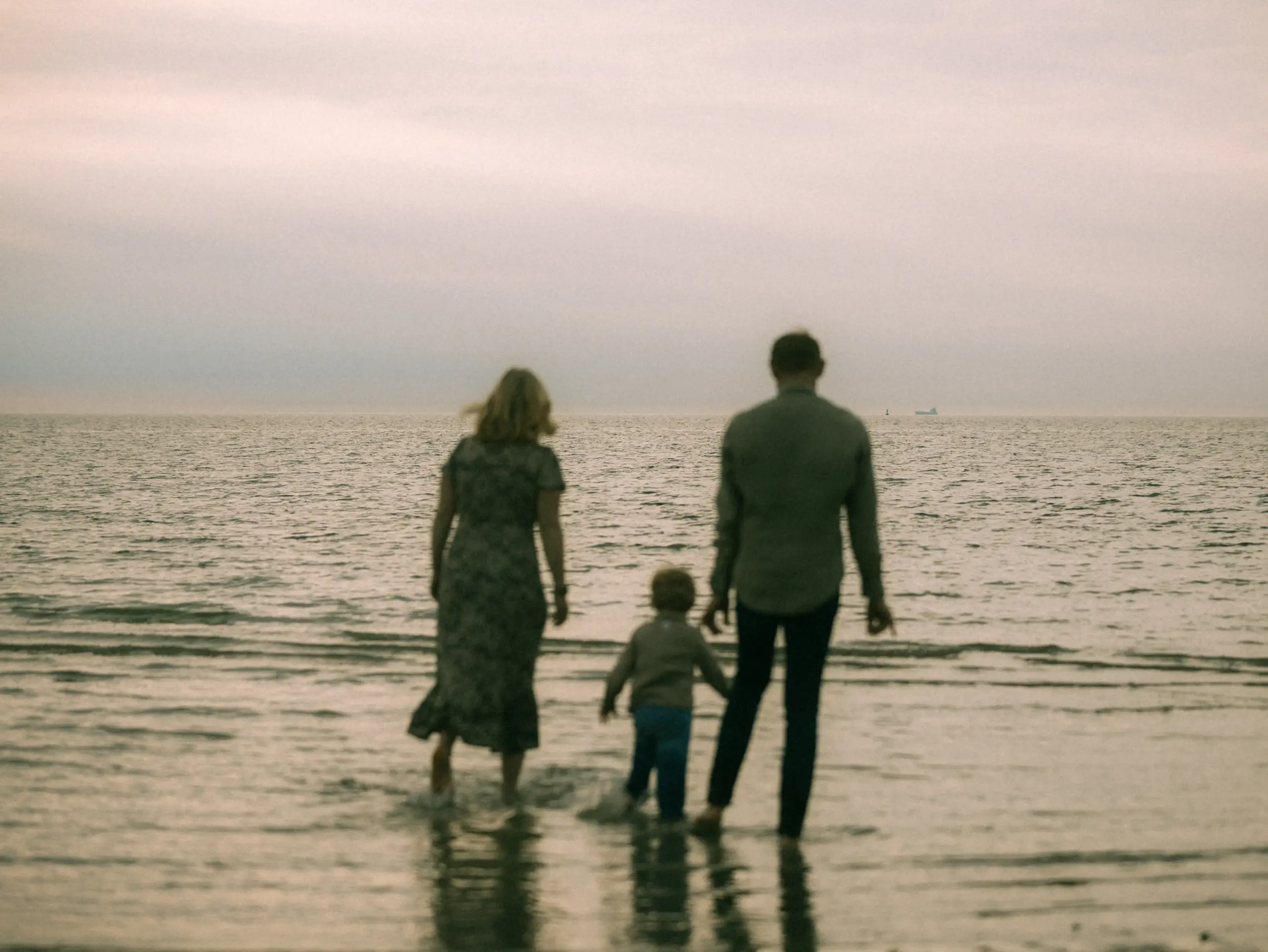 Eine junge Familie steht gemeinsam im flachen Wasser am Meer bei bewölktem Himmel. Die Eltern halten ihr kleines Kind an den Händen, während sie in Richtung Horizont blicken. Stimmungsvolle Familienfotos in Schwerin, die eine intime und natürliche Bindung am Strand zeigen.