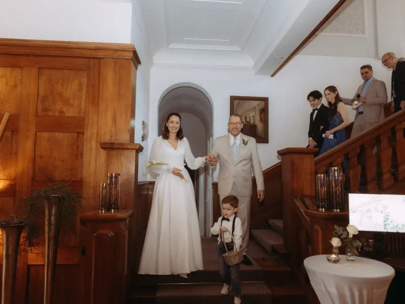 Ein Brautpaar schreitet glücklich die Holztreppe hinab, begleitet von einem kleinen Jungen mit Blumenkorb. Das elegante Ambiente des historischen Gebäudes unterstreicht diesen besonderen Moment, festgehalten von einem Hochzeitsfotograf in Schwerin und MV.
