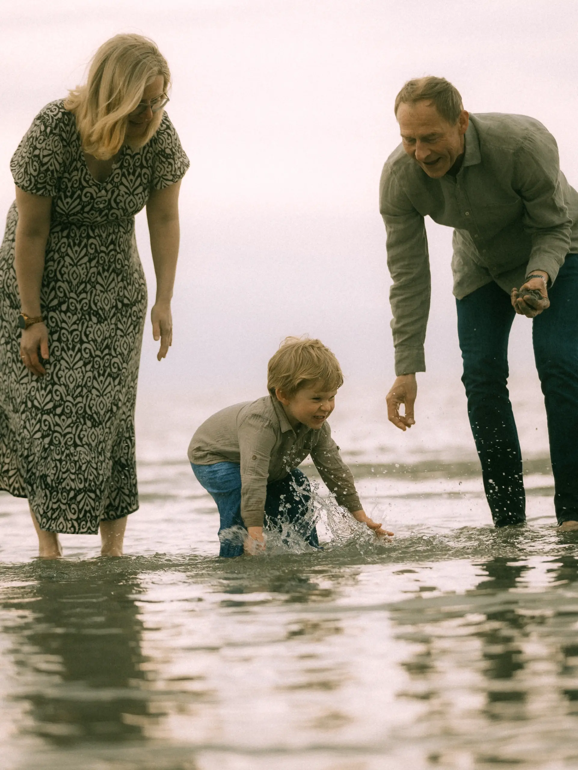 Ein glücklicher Junge spielt am Strand im flachen Wasser, während seine Eltern daneben stehen und zuschauen. Die Familie genießt einen entspannten Tag am Meer. Diese Familienfotos in Schwerin halten solch natürliche und freudige Momente fest.